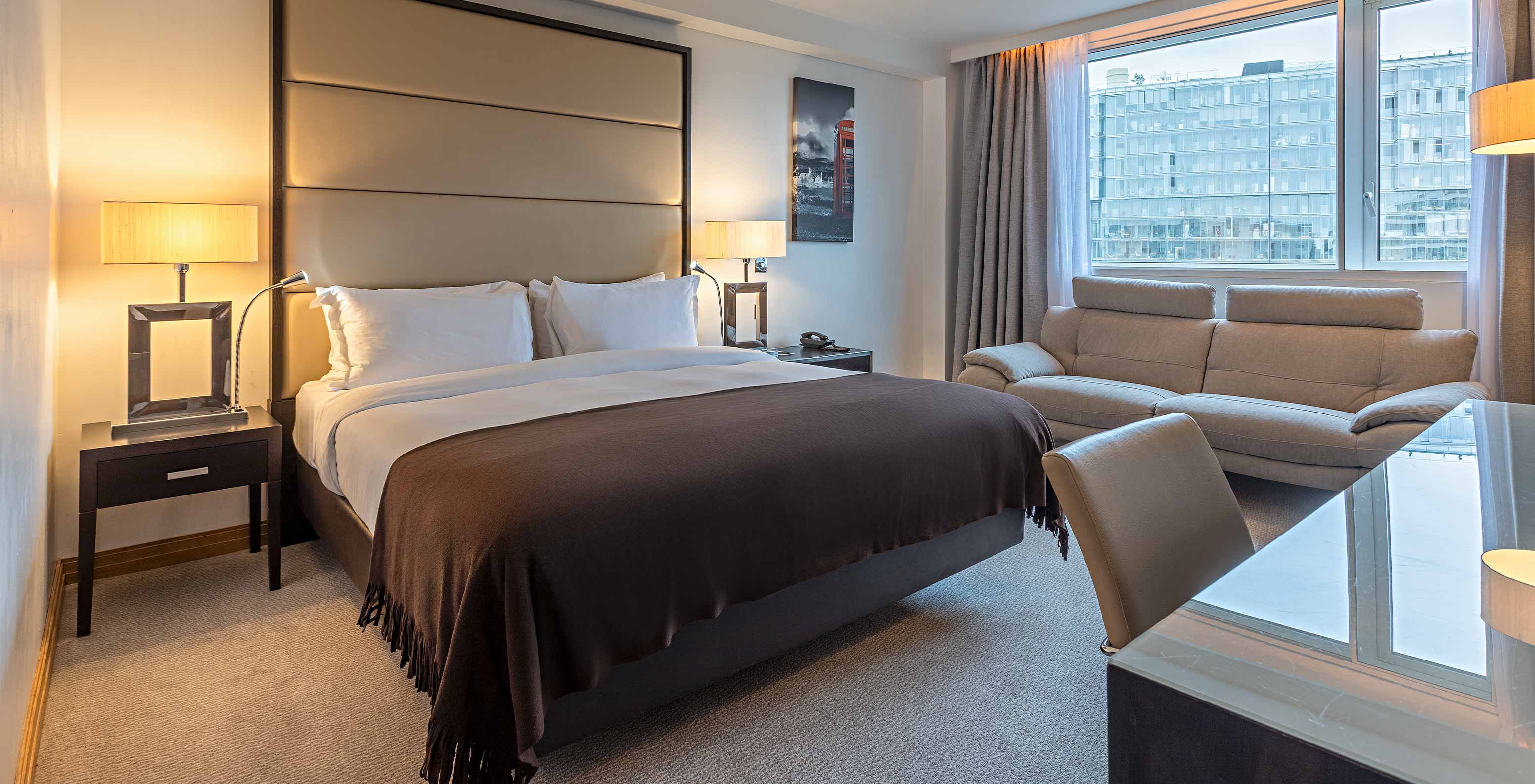 Dormitorio moderno con cama grande, sofá junto a la ventana y escritorio iluminado con vista urbana