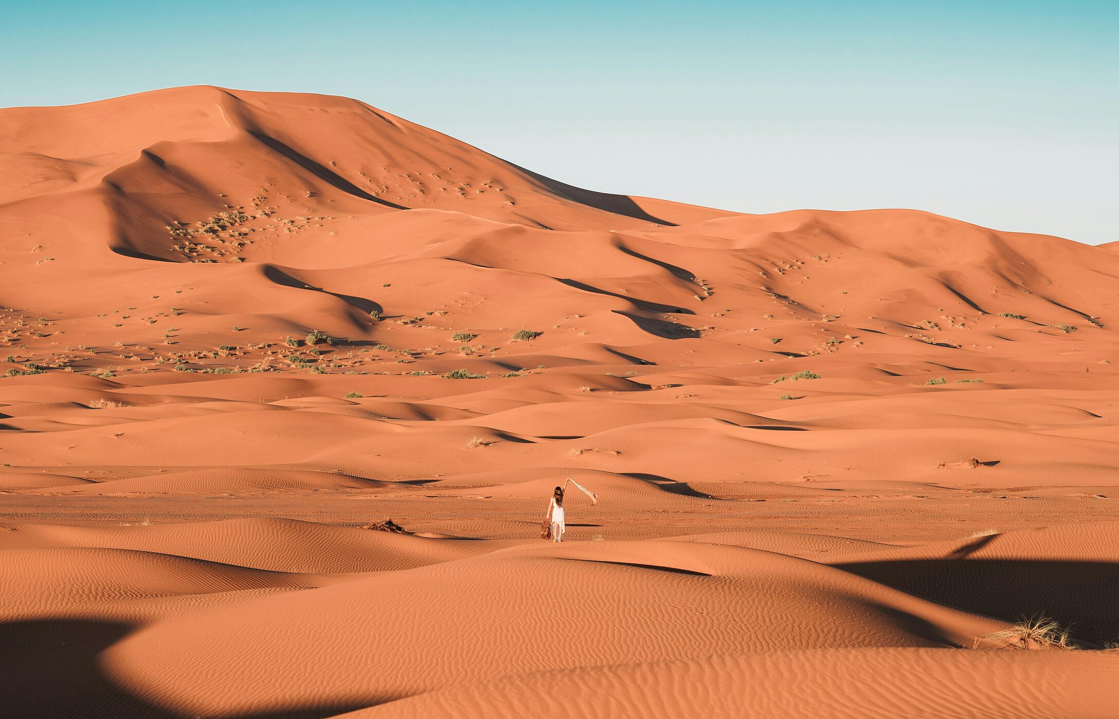 Femme solitaire, vêtue de blanc, au milieu des dunes du désert du Sahara au Maroc, avec quelques végétations en arrière-plan.