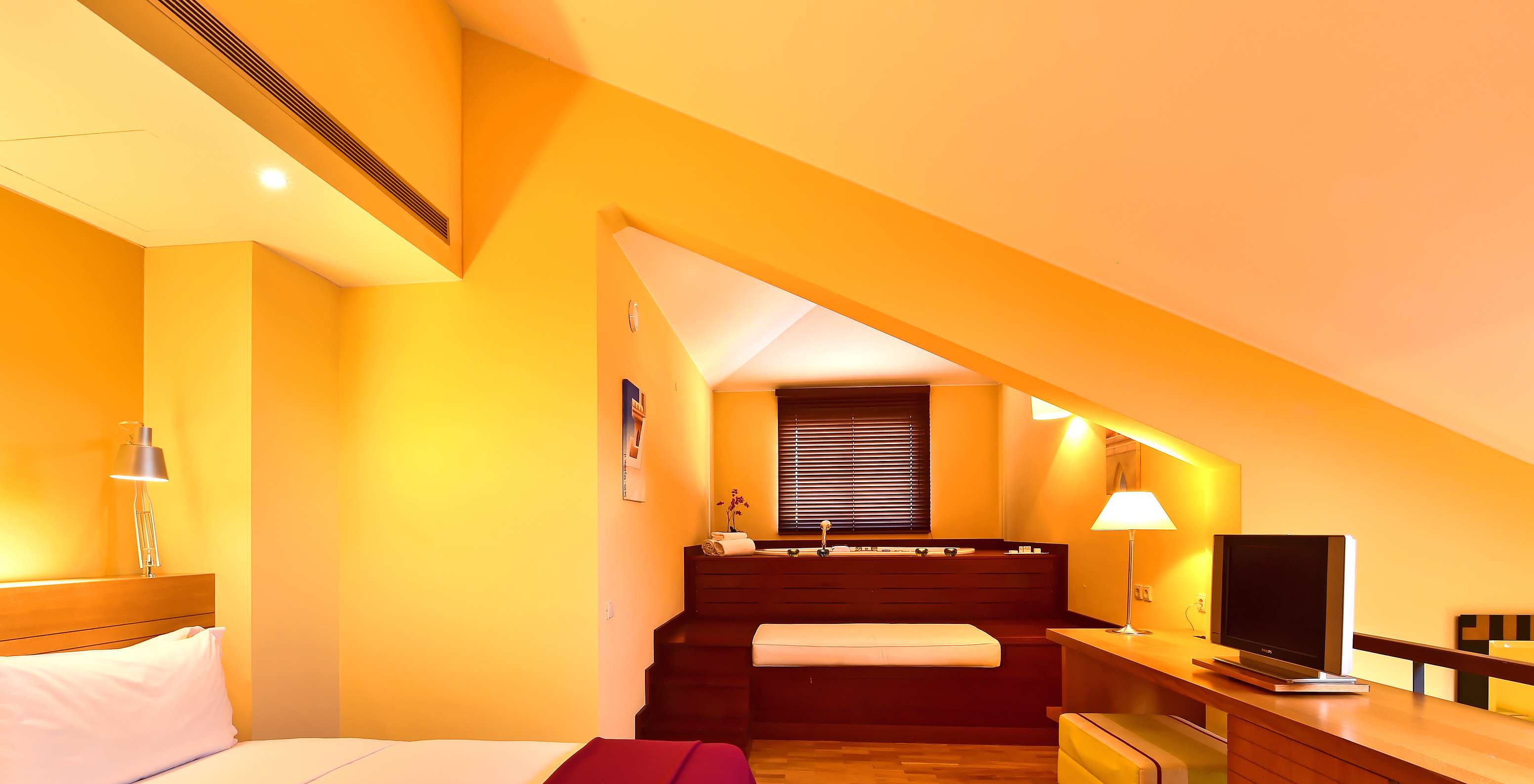 Chambre mansardée aux tons chauds avec lit, baignoire intégrée en bois, bureau, lampe et télévision