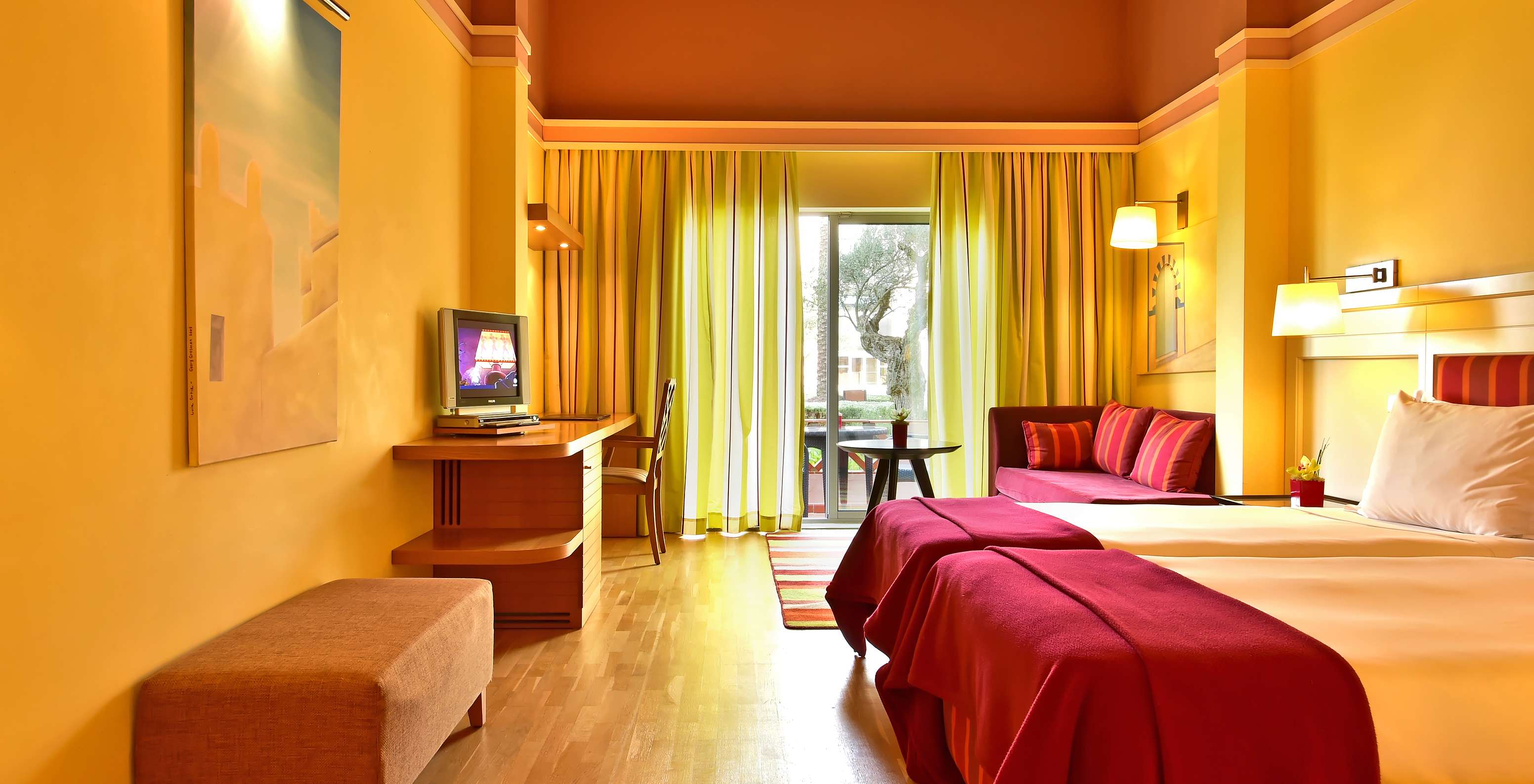 Chambre lumineuse avec deux lits, canapé rouge, bureau en bois et baie vitrée donnant sur un balcon
