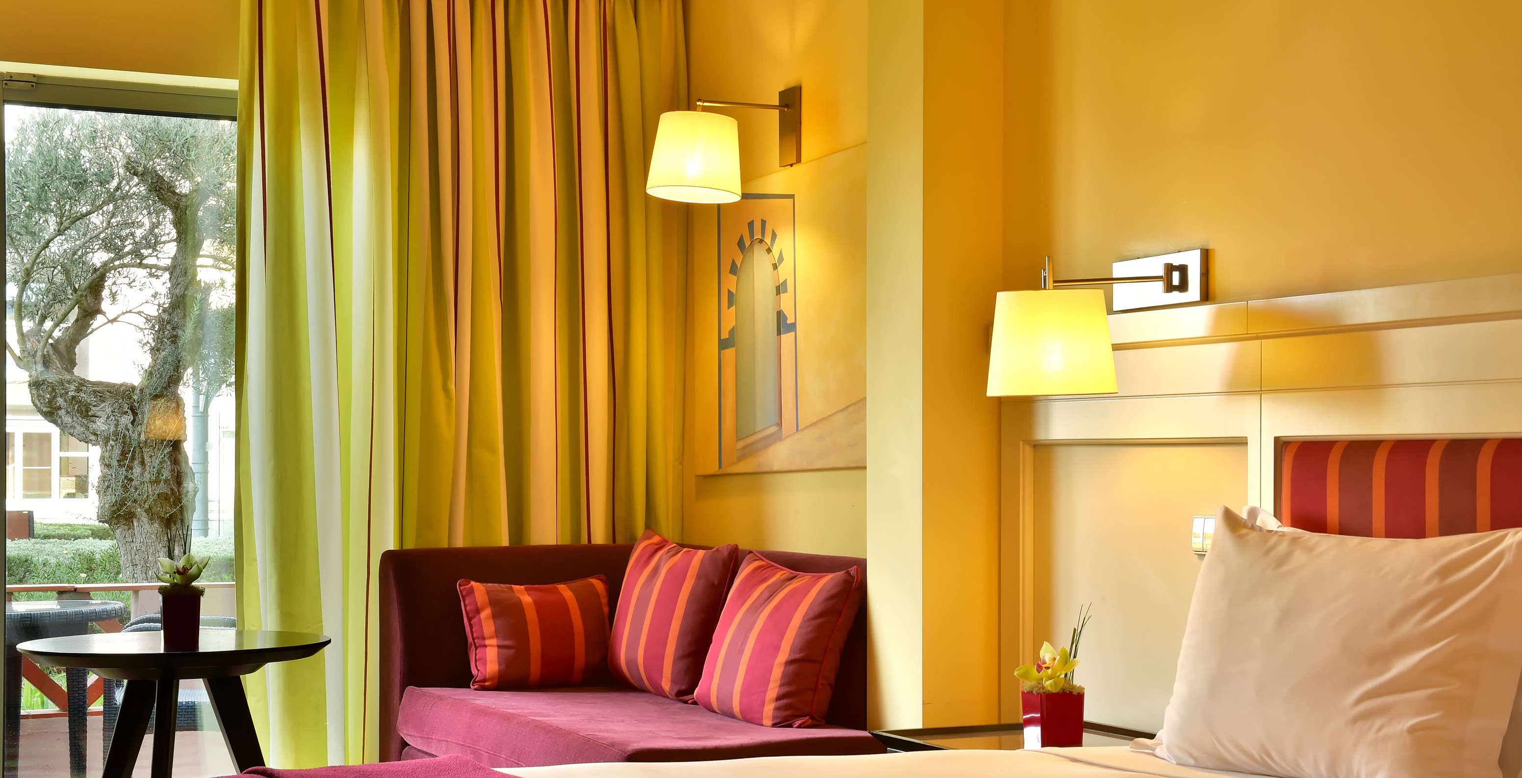 Chambre lumineuse avec canapé rouge, rideaux colorés, lampes murales et grande fenêtre sur un arbre