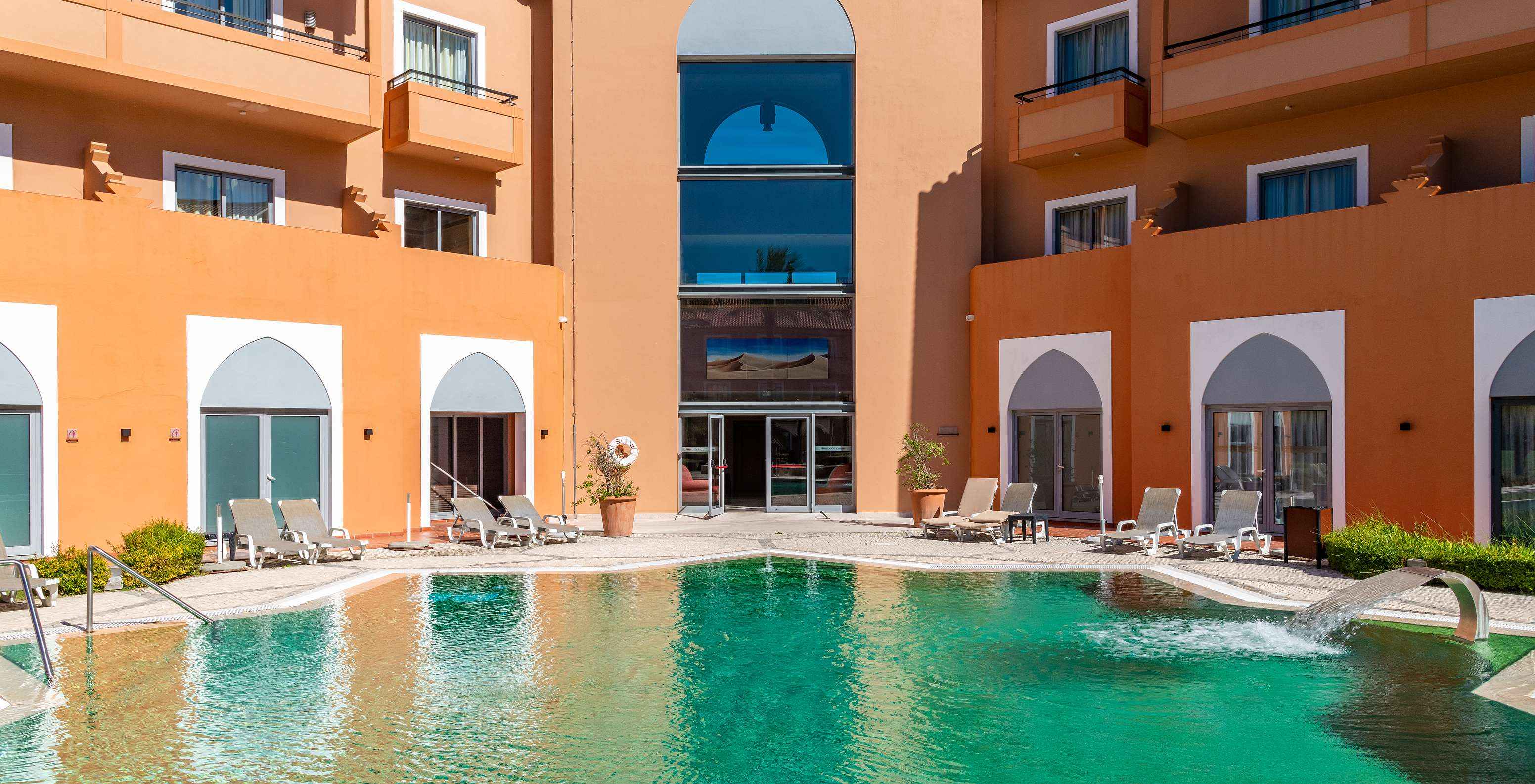 Piscine extérieure entourée de transats devant la façade ocre de l’hôtel avec balcons et grandes baies vitrées