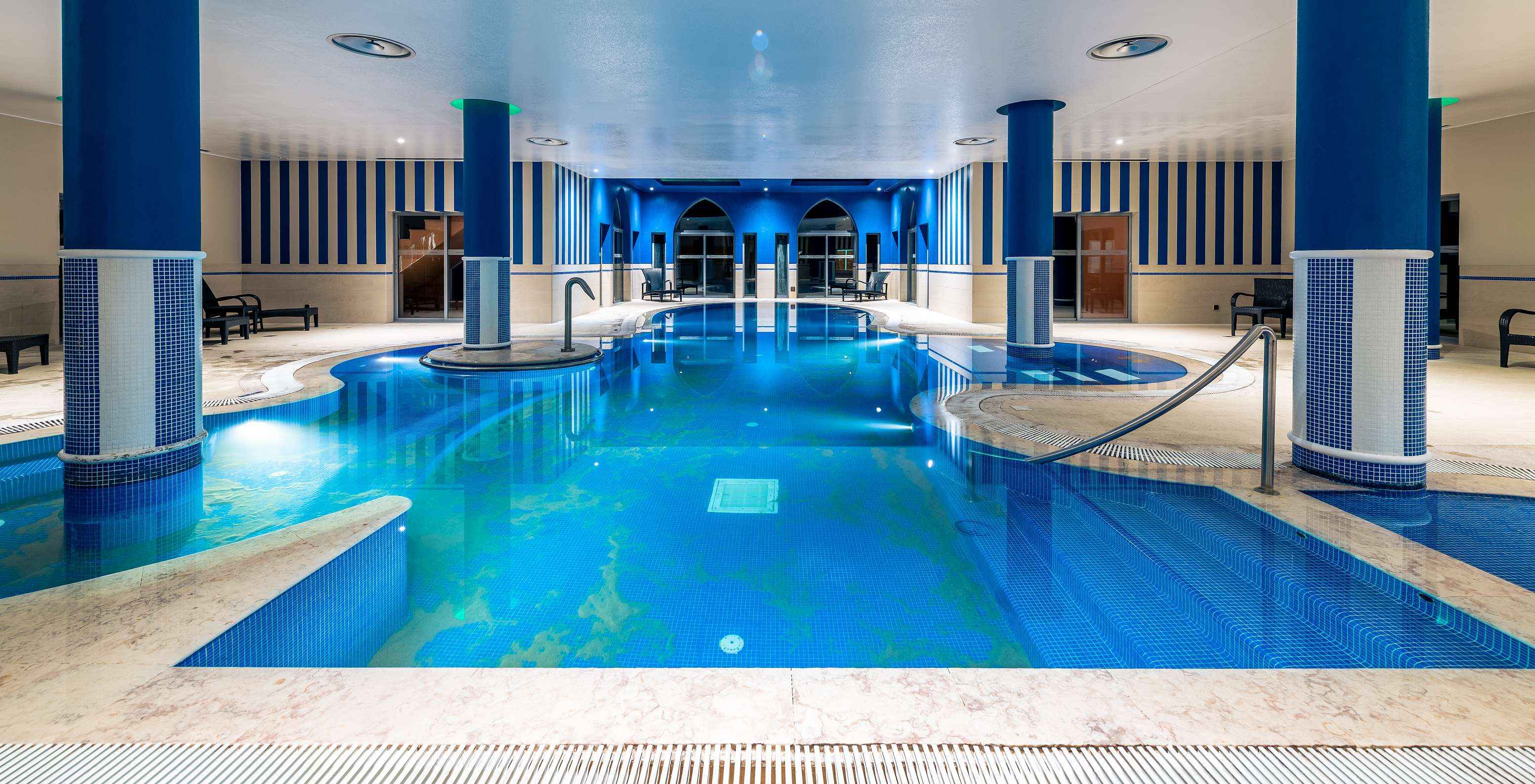 Piscine intérieure aux tons bleus avec colonnes carrelées, éclairage tamisé et espaces de détente autour
