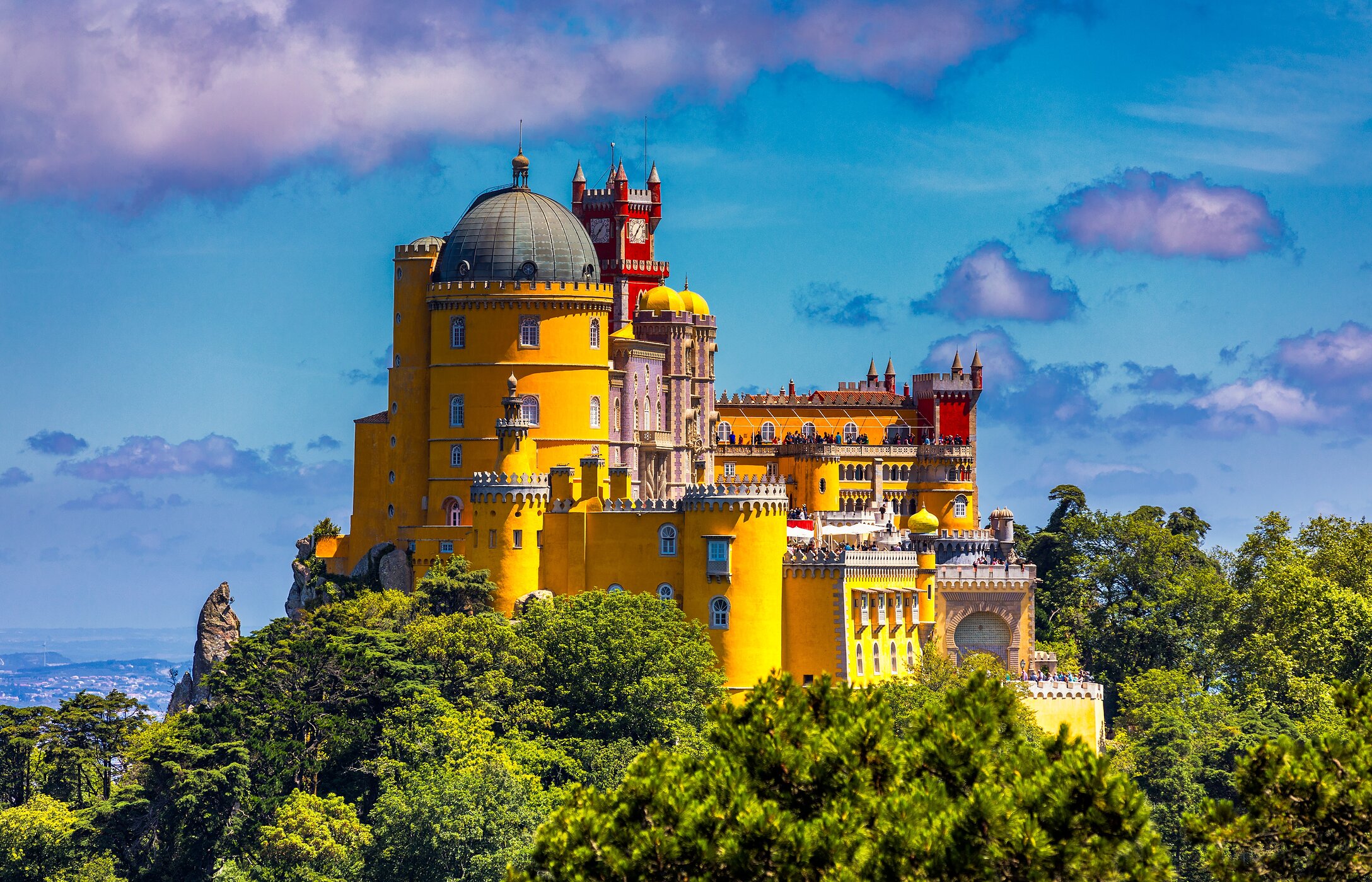 Vue aérienne du Palais de Pena à Sintra, avec ses couleurs vives, entouré de végétation sous un ciel bleu.