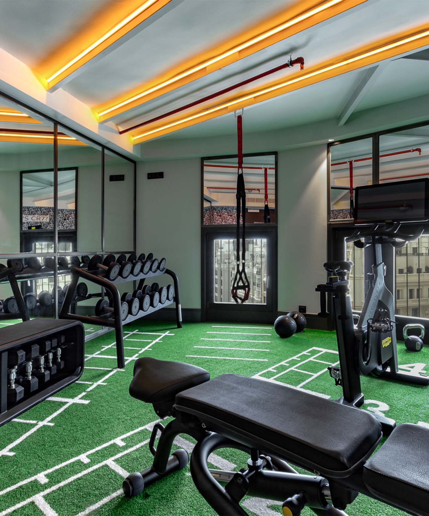 Salle de sport avec haltères, vélo, banc de musculation et TRX, avec miroirs et grandes fenêtres avec vue sur les bâtiments
