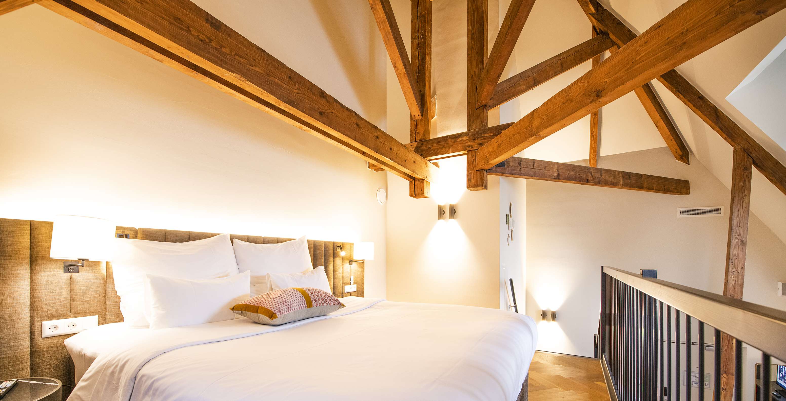 Chambre mansardée avec grand lit blanc, poutres apparentes en bois et éclairage chaleureux sous les combles