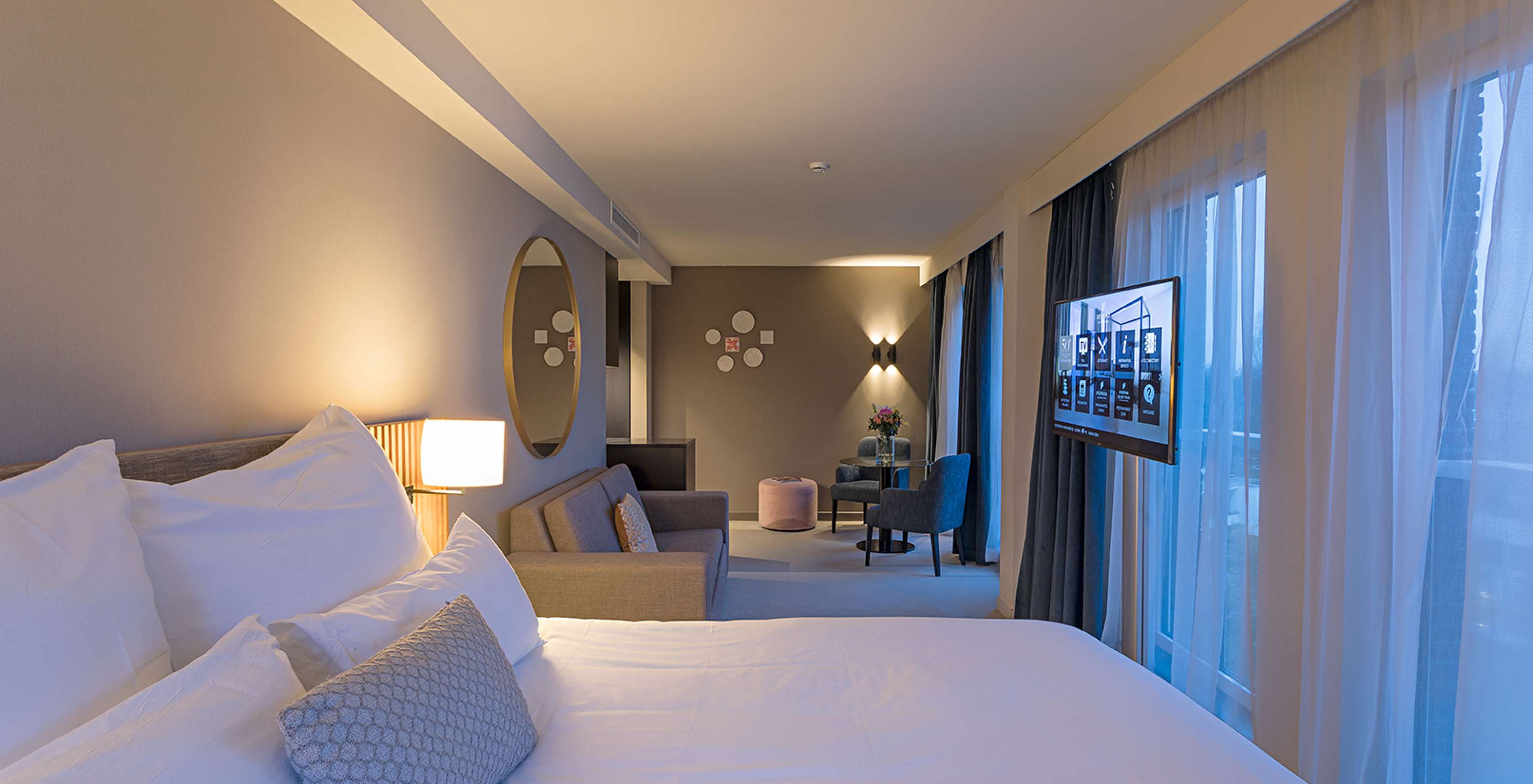 Chambre d’hôtel lumineuse avec grand lit, coin salon moderne, télévision murale et fenêtres voilées bleues