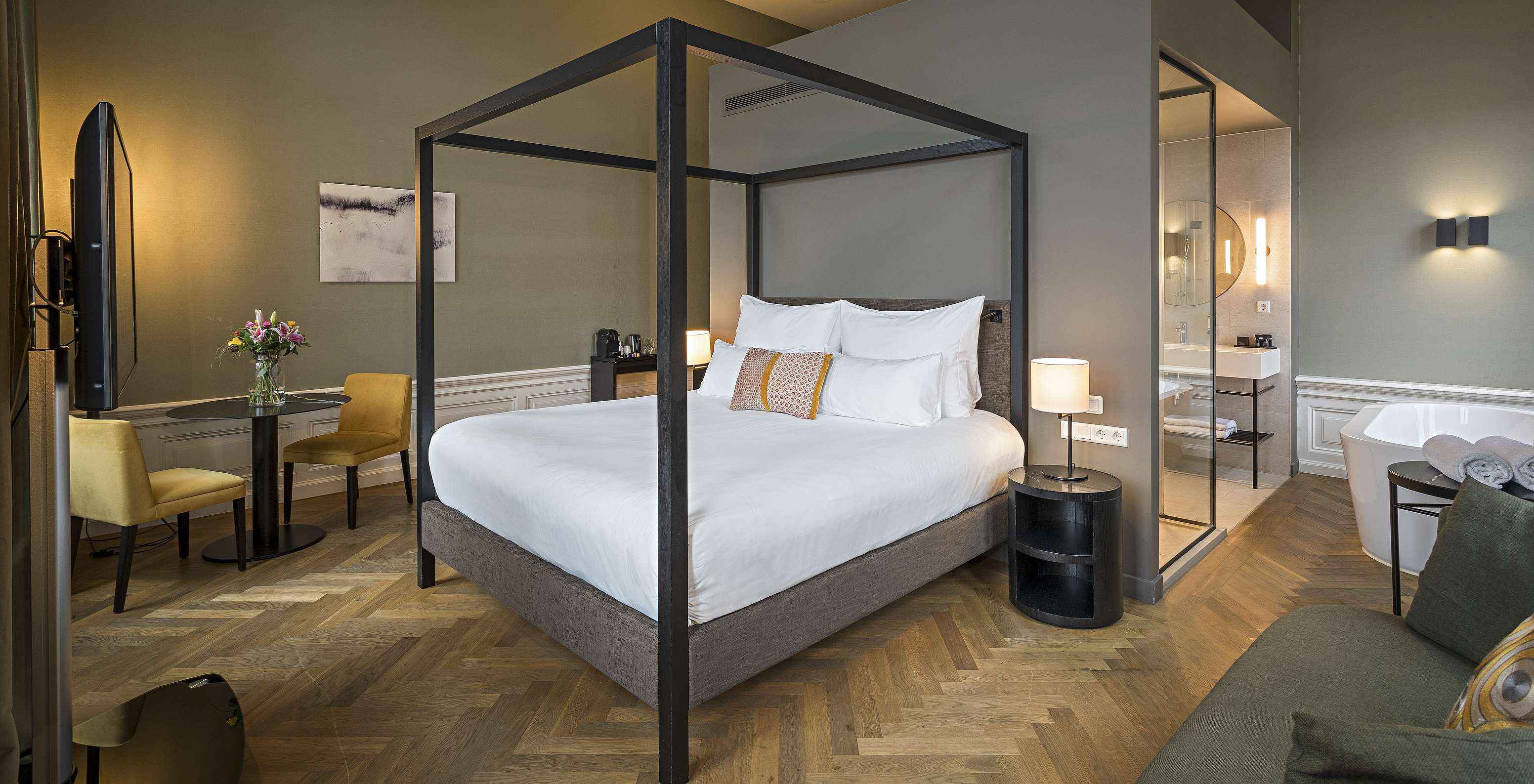 Chambre élégante avec lit à baldaquin, coin salon, table ronde et salle de bain ouverte aux tons sobres