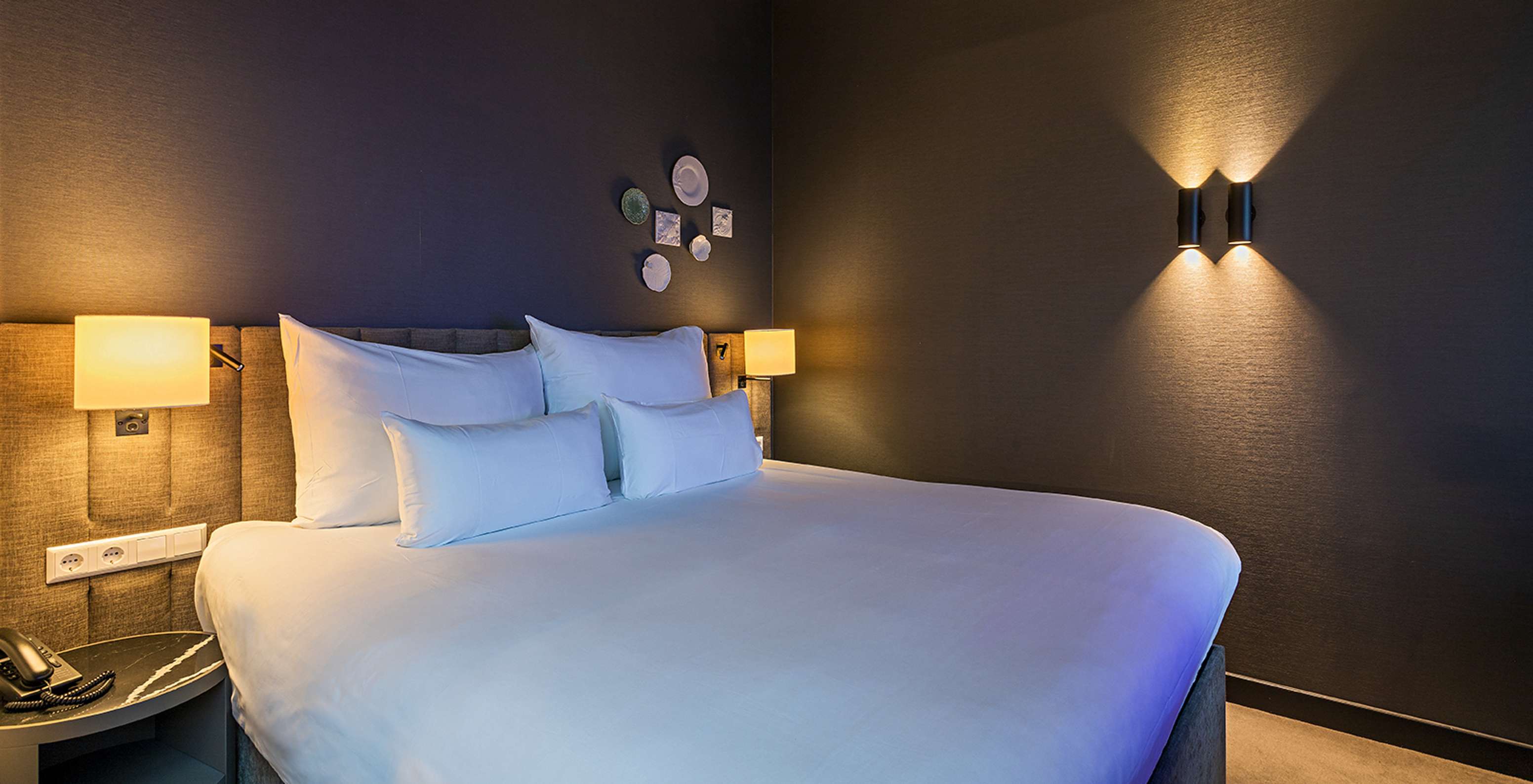 Grand lit blanc dans une chambre sombre avec lampes murales allumées et tête de lit capitonnée aux tons bruns