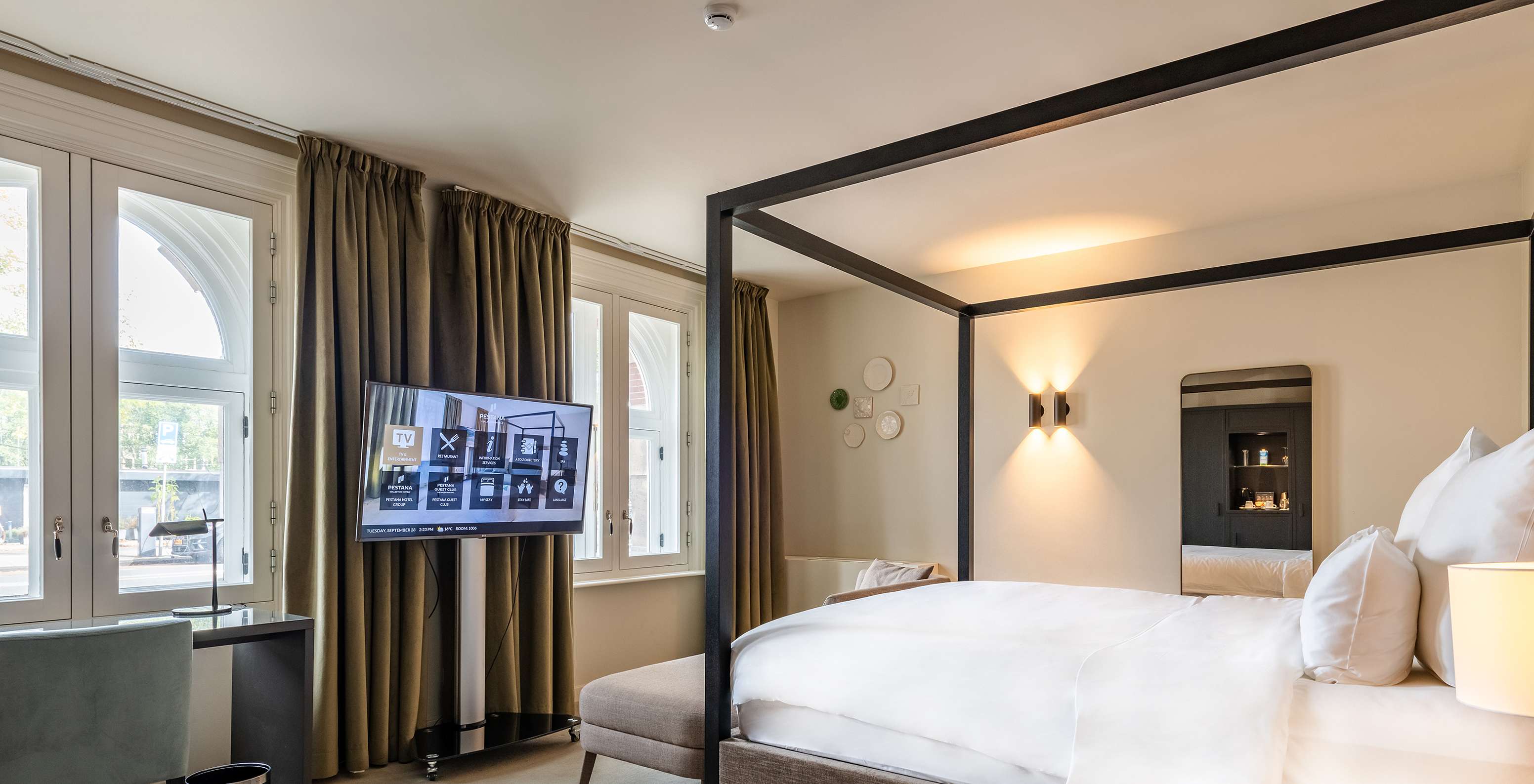 Chambre lumineuse avec lit à baldaquin, télévision sur pied, grandes fenêtres et rideaux sombres