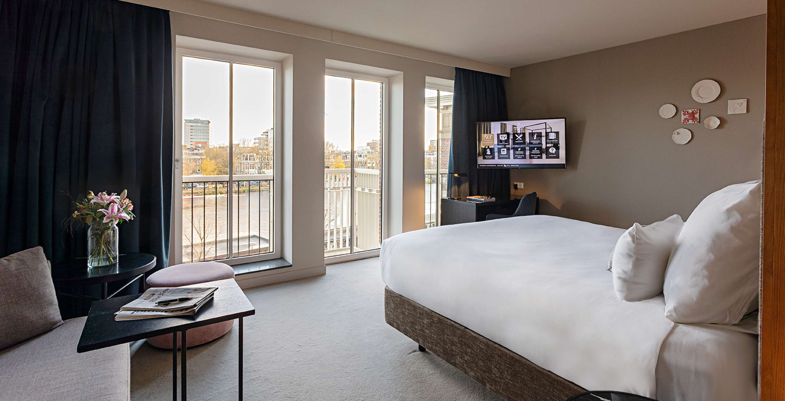 Chambre lumineuse avec grand lit, télévision murale et vue sur le canal à travers de hautes fenêtres