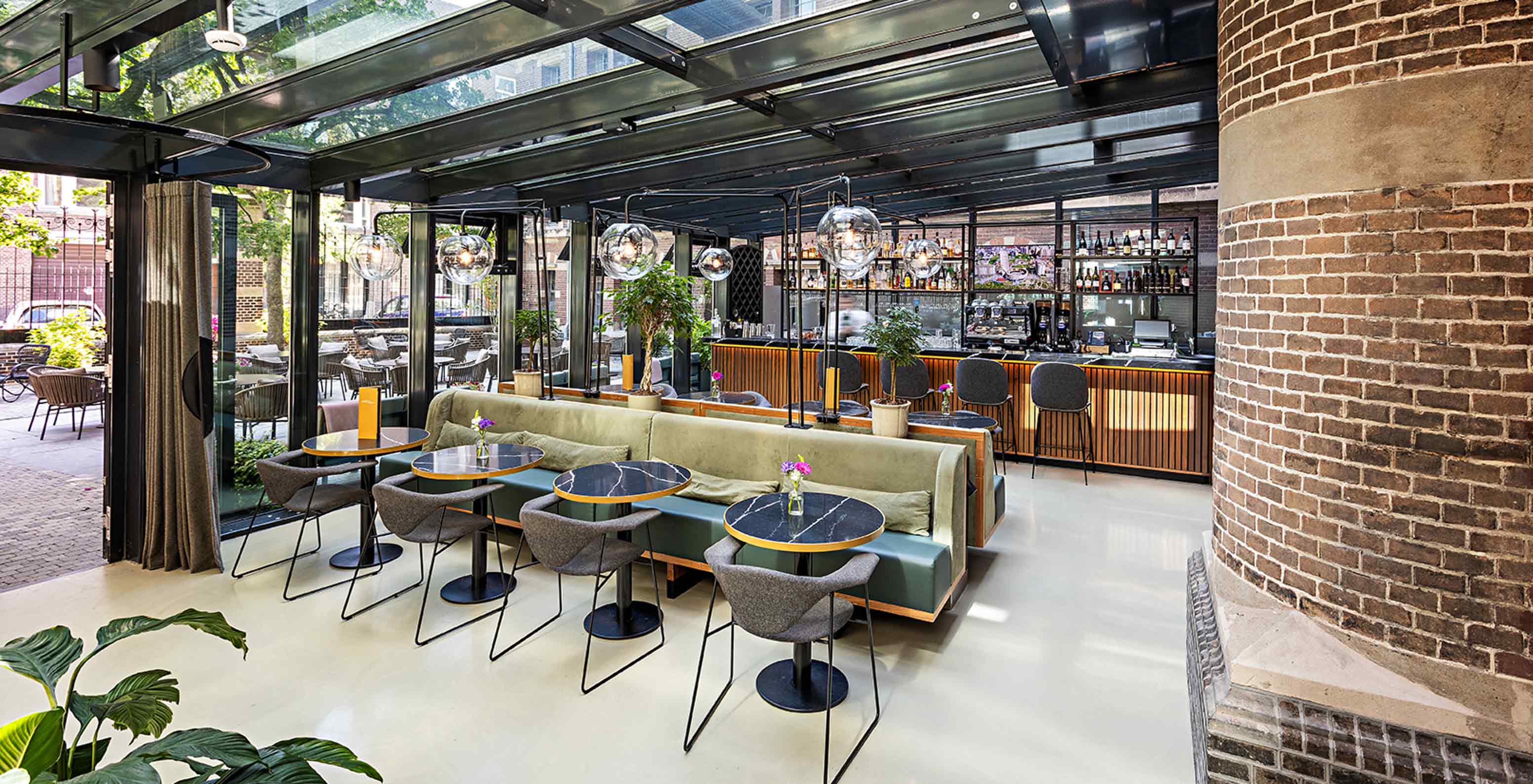 Espace bar et restaurant lumineux avec banquettes vertes, tables rondes et grande verrière ouverte