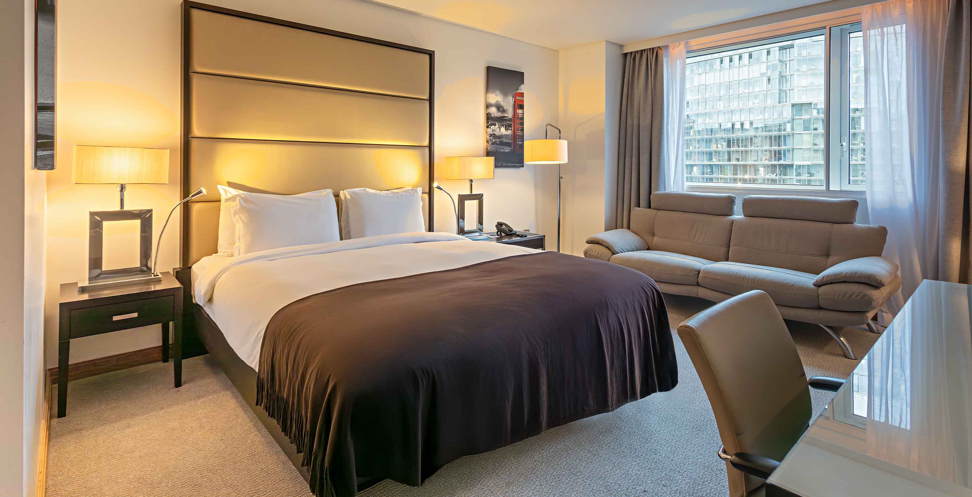 Chambre moderne avec grand lit, canapé gris, bureau brillant et large fenêtre sur des immeubles urbains