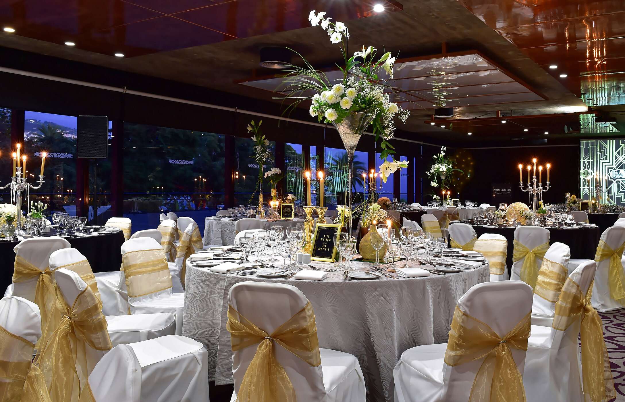 Mariage dans une salle moderne avec tables rondes, arrangements floraux hauts et éclairage doux et élégant