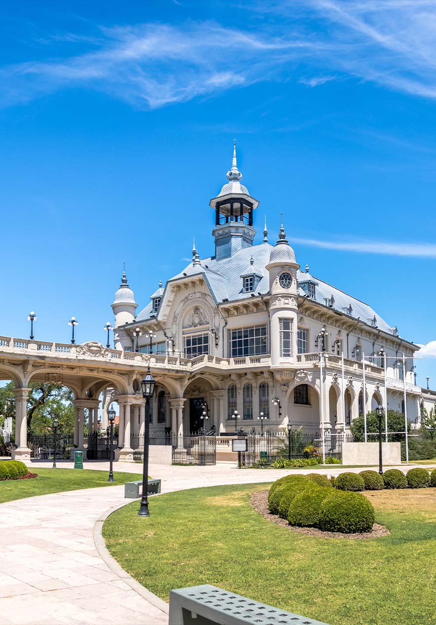 Palácio histórico em Buenos Aires com torres ornamentadas, jardins verdes e céu azul com nuvens leves