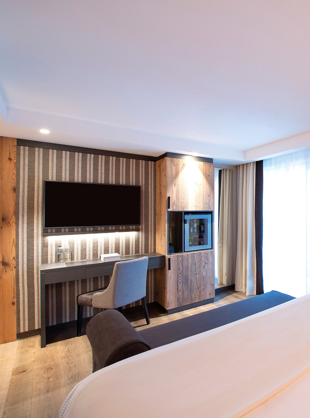 Quarto Deluxe no Le Massif Courmayeur com cama ampla, mesa de trabalho, TV na parede e cortinas claras