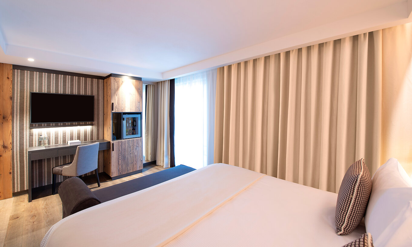Quarto Grande Deluxe do Le Massif Courmayeur com cama ampla, mesa de trabalho, TV e cortinas iluminadas