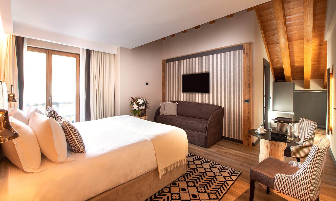Suíte Júnior do Le Massif Courmayeur com cama ampla, sofá escuro, mesa lateral e teto de madeira claro