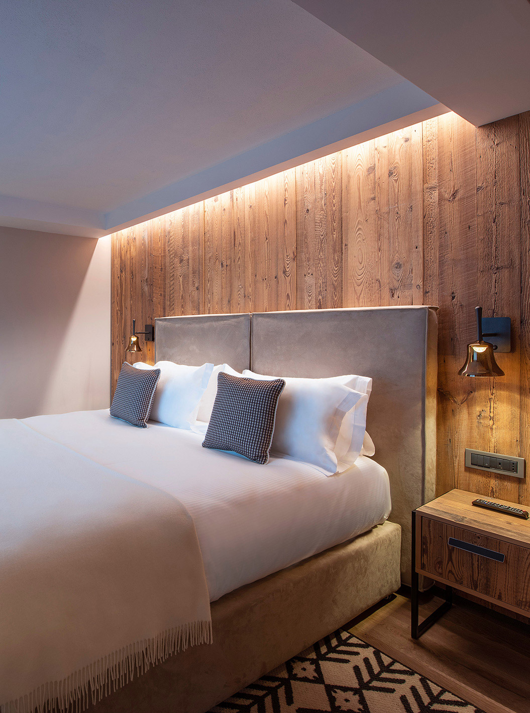 Suíte do Le Massif Courmayeur com cama ampla, painel de madeira iluminado, TV na parede e tapete geométrico