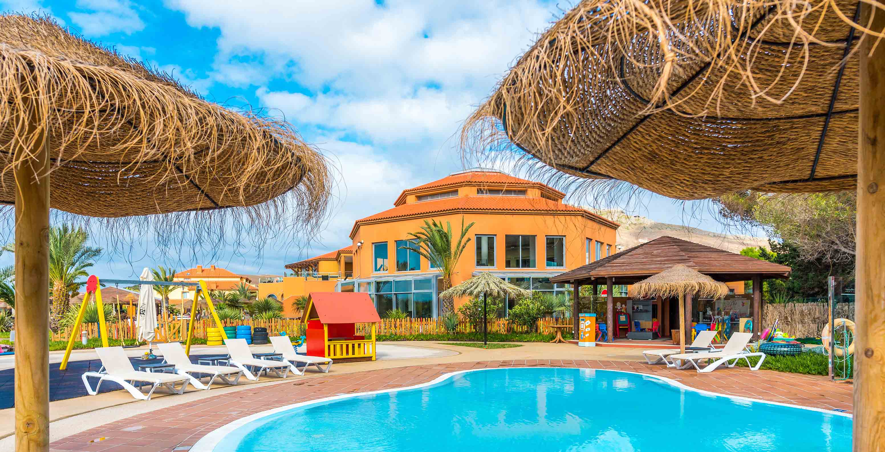 Piscina infantil e parque de jogos coloridos no Pestana Porto Santo, com chapéus de sol de palha e um edifício ao fundo