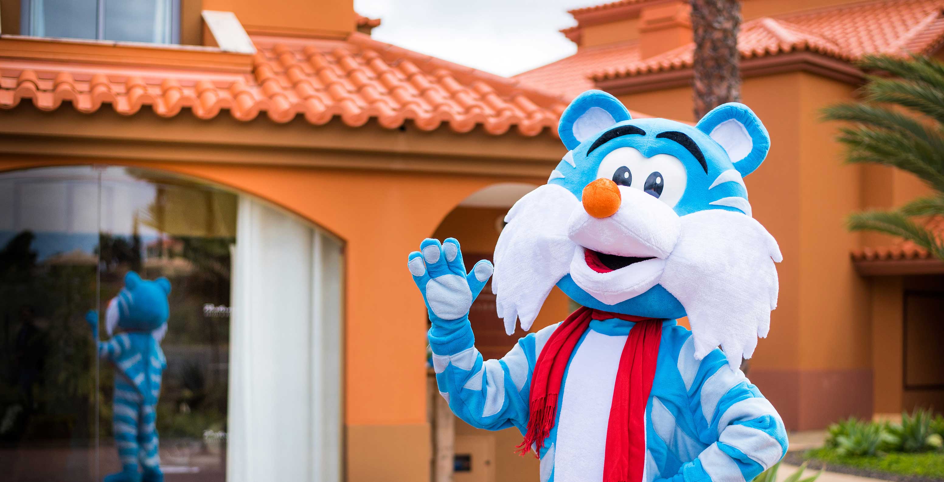 Mascote tigre azul e branco acenando em frente ao Pestana Porto Santo, com reflexo visível na janela