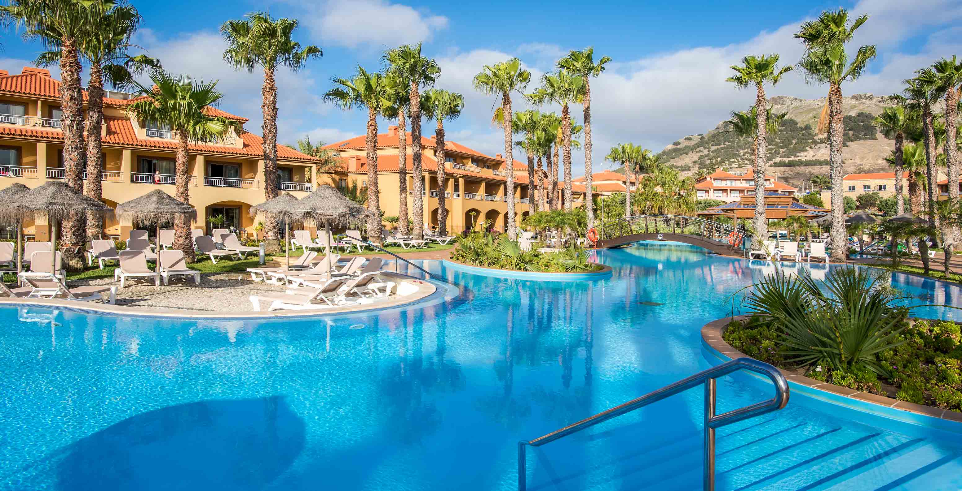 Vista da piscina do Pestana Porto Santo rodeada por palmeiras e edifícios coloridos sob um céu azul