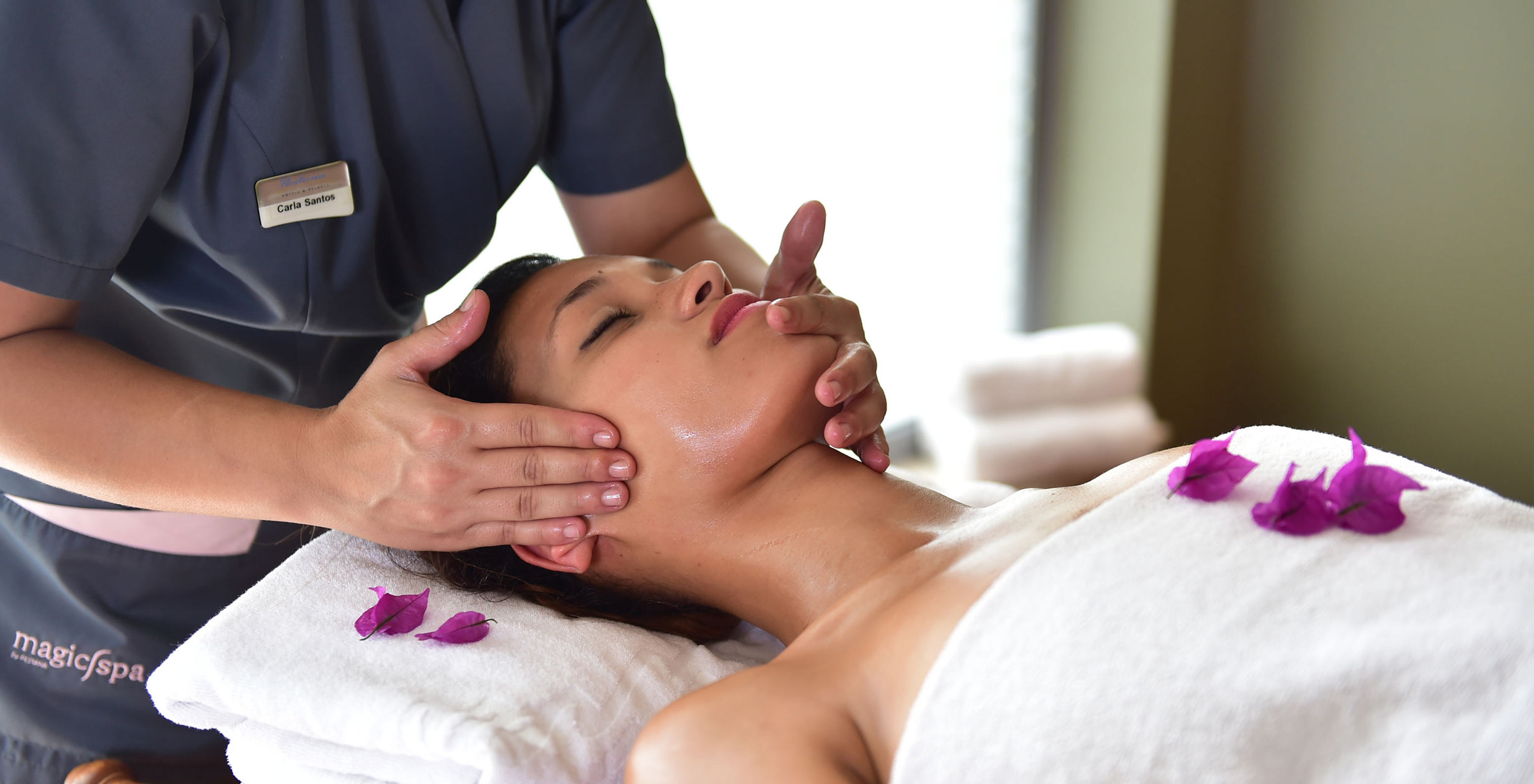 Mulher a receber uma massagem facial no Pestana Porto Santo, com pétalas roxas e mãos de terapeuta visíveis