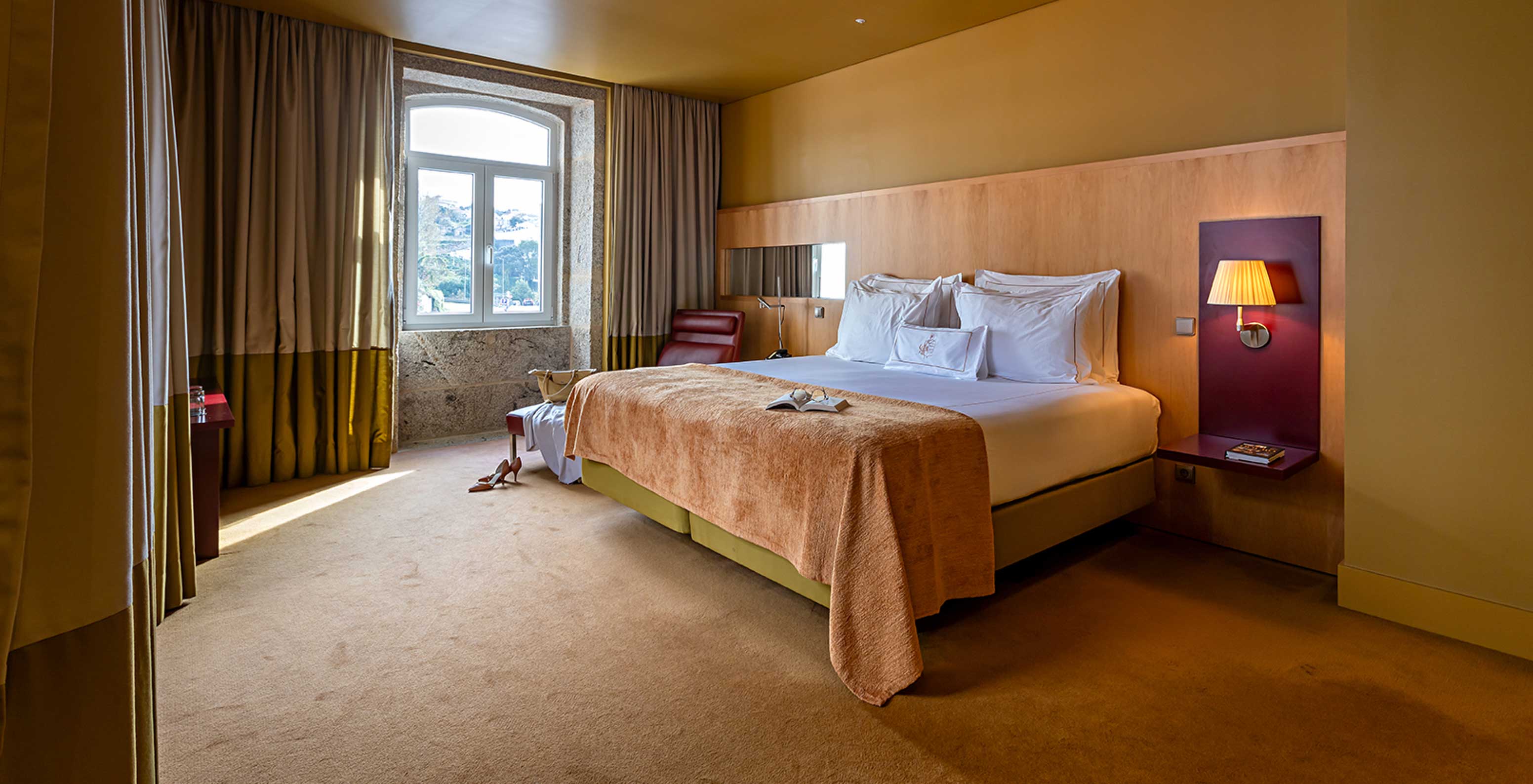A Junior Suite do Pestana Palácio do Freixo tem uma cama de casal com uma manta beje e uma janela em pedra com vista rio
