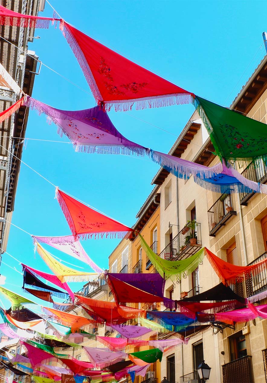 Lenços coloridos suspensos entre prédios históricos iluminam uma rua estreita sob céu azul em Madri