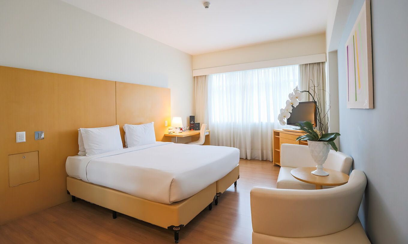 O Queen Superior do Pestana Curitiba tem um quarto com cama de casal, sofás, uma secretária e um móvel com televisão