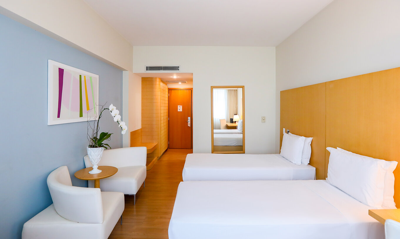 O Twin Superior do Pestana Curitiba tem duas camas individuais com sofás nos pés da cama e uma pequena mesa com flores