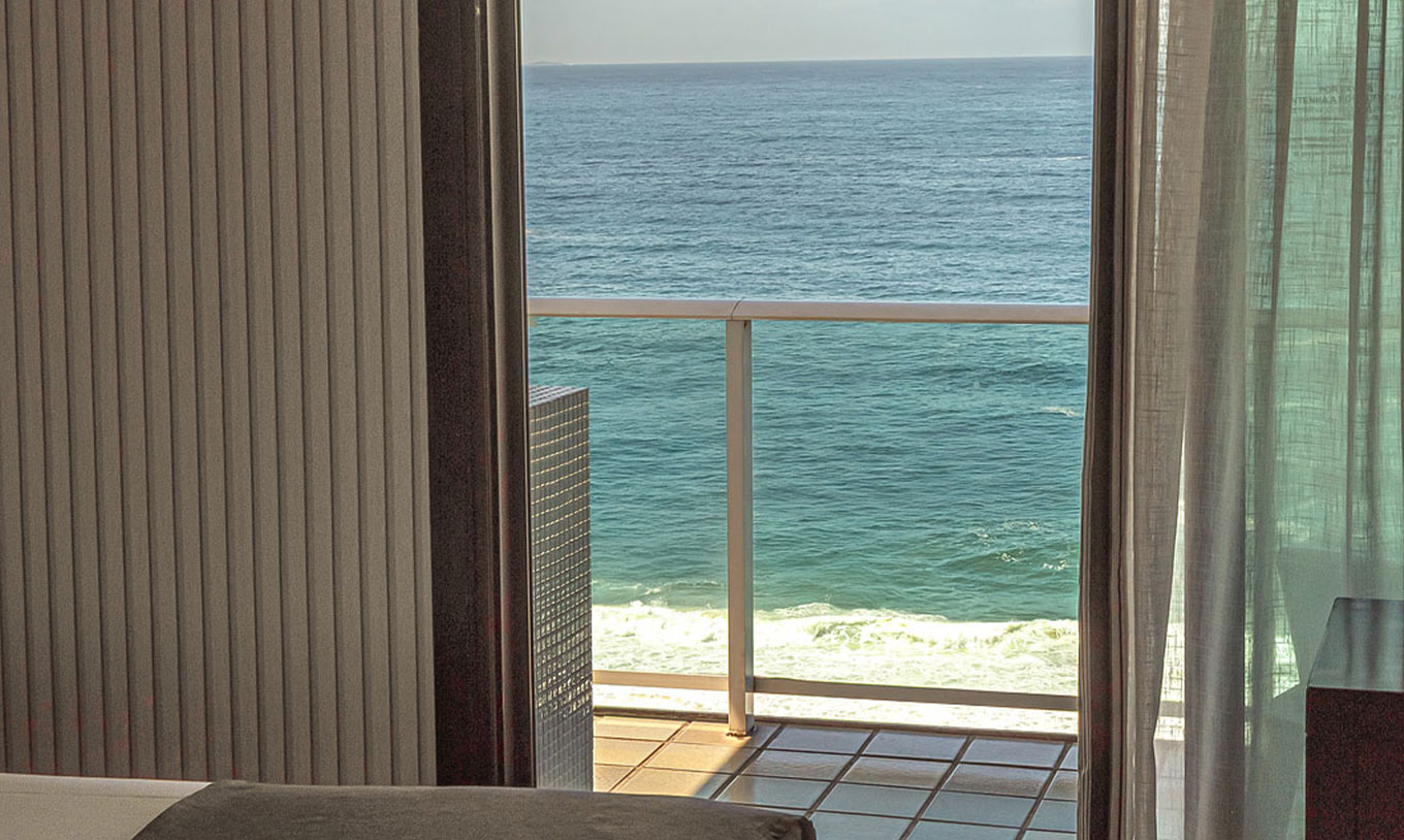 A Suite Frente Praia do Pestana Rio Atlântica tem uma varanda com cortinas abertas e vista para o mar a partir do quarto