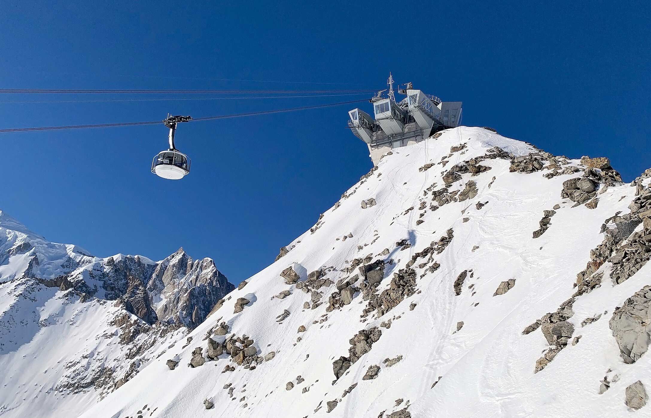 Teleférico suspenso junto a uma estação no topo nevado da montanha sob céu azul intenso e picos rochosos