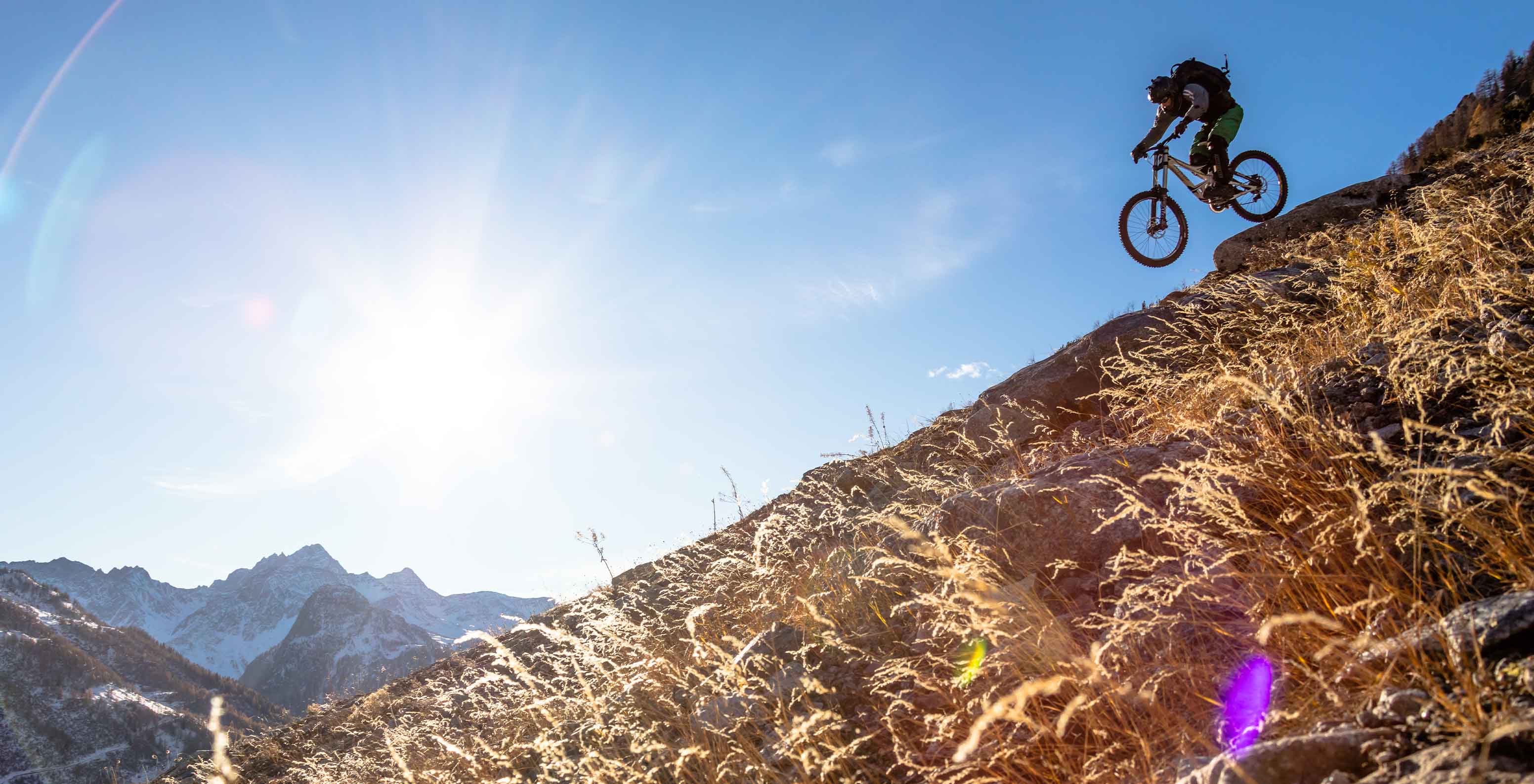 Ciclista a descer trilho de montanha perto do Le Massif Courmayeur, com sol e picos nevados ao fundo