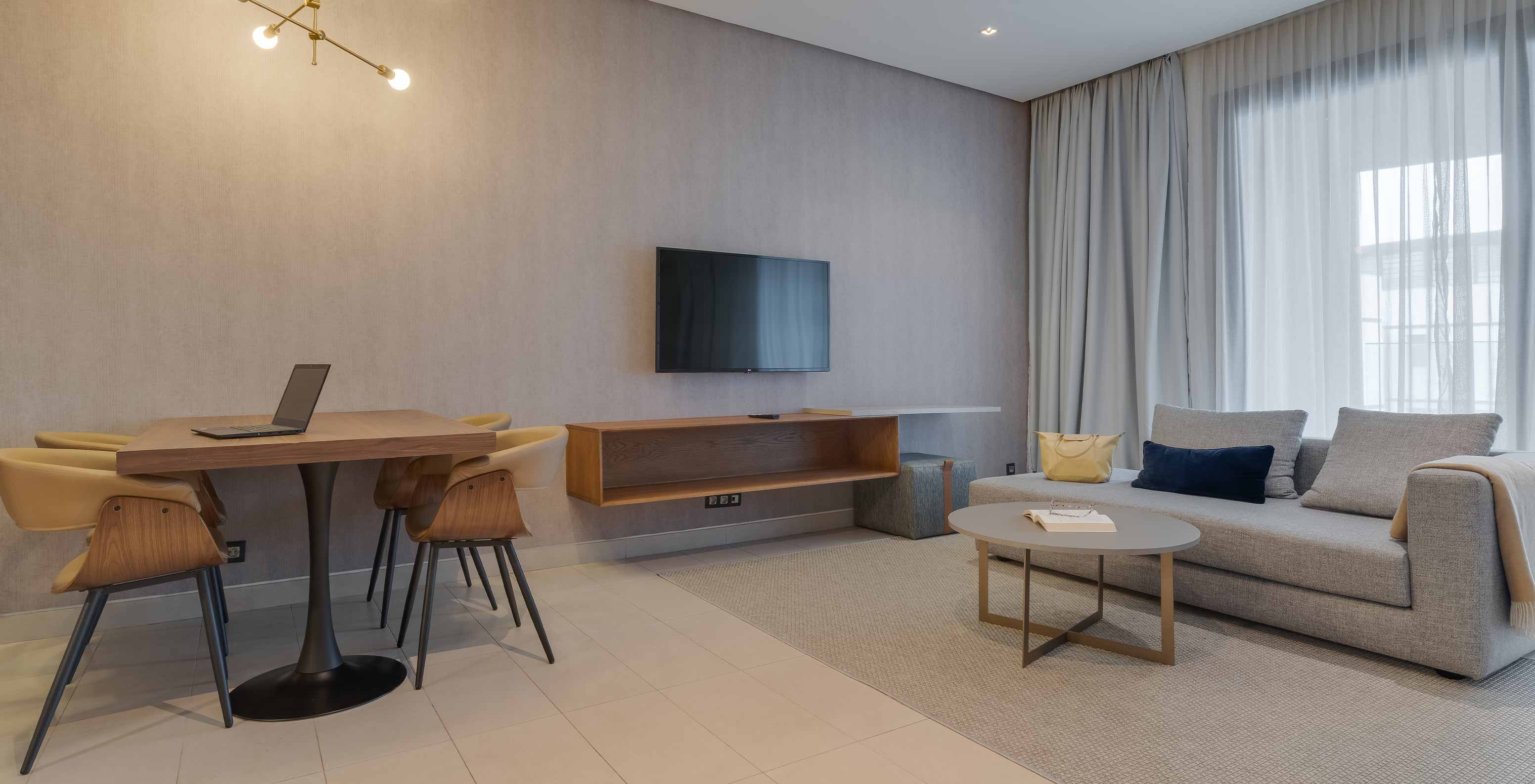Suite Júnior do Pestana Casablanca com sofá cinzento, mesa redonda, televisão e cortinas amplas junto à janela