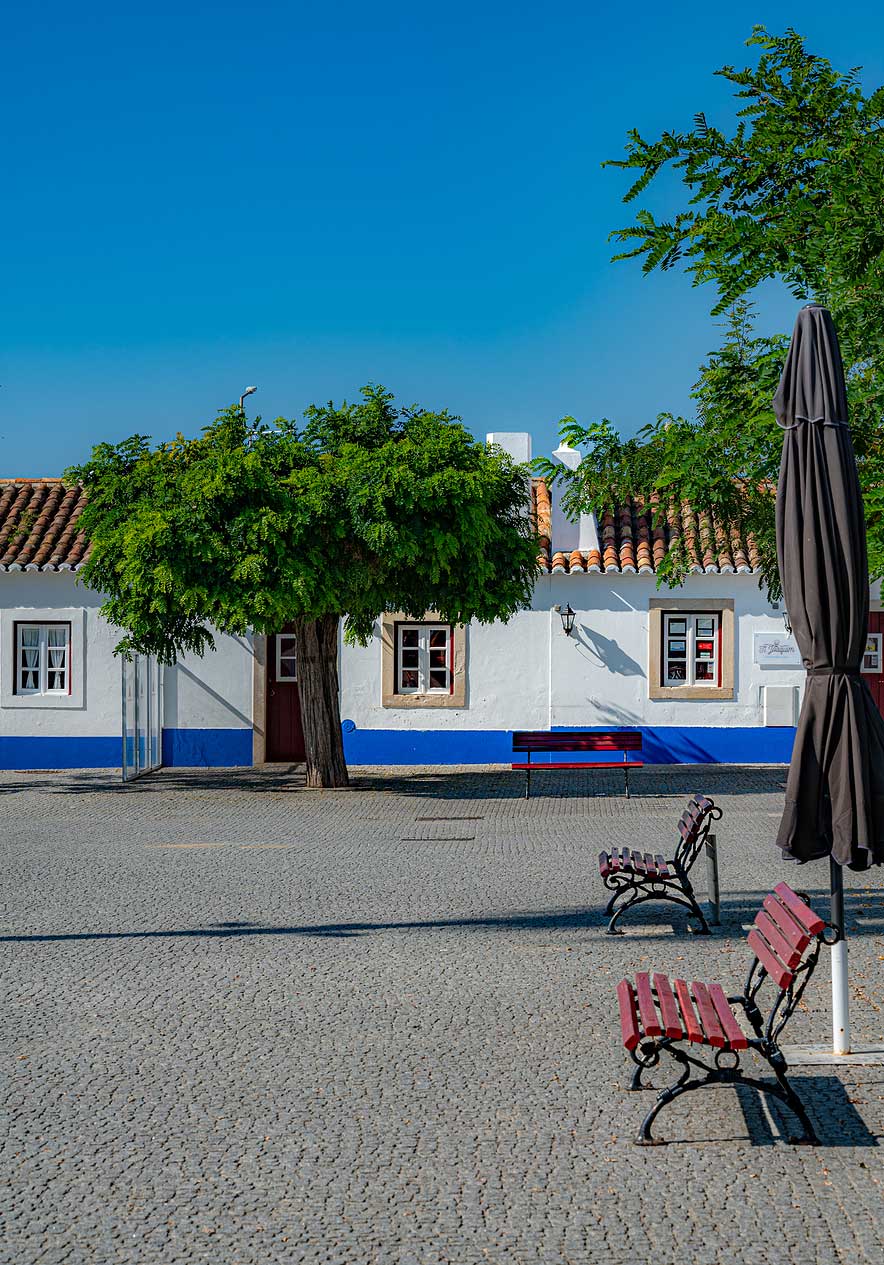 Praça com bancos vermelhos e casas brancas de faixa azul no centro histórico perto do Pestana Porto Covo Village