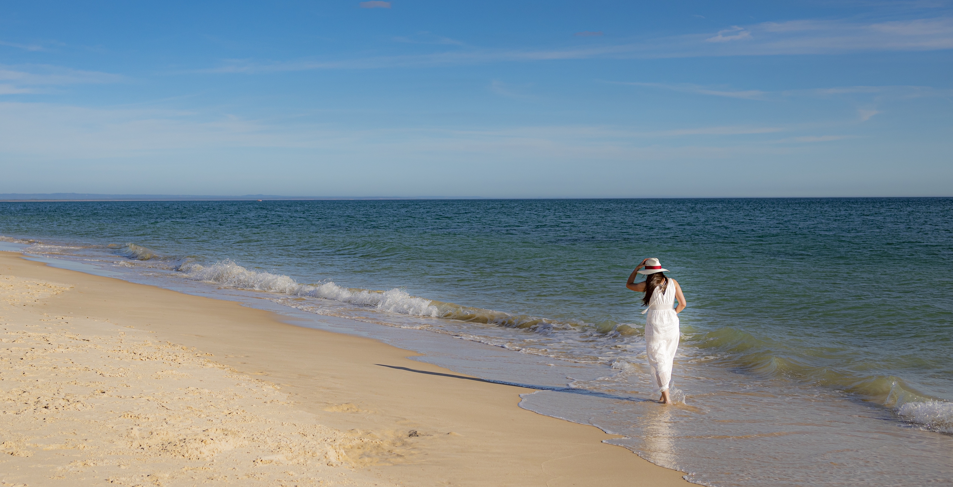 Mulher com vestido e chapéu branco passeia à beira-mar ao pé do Pestana Tróia Eco-Resort, com acesso direto à praia