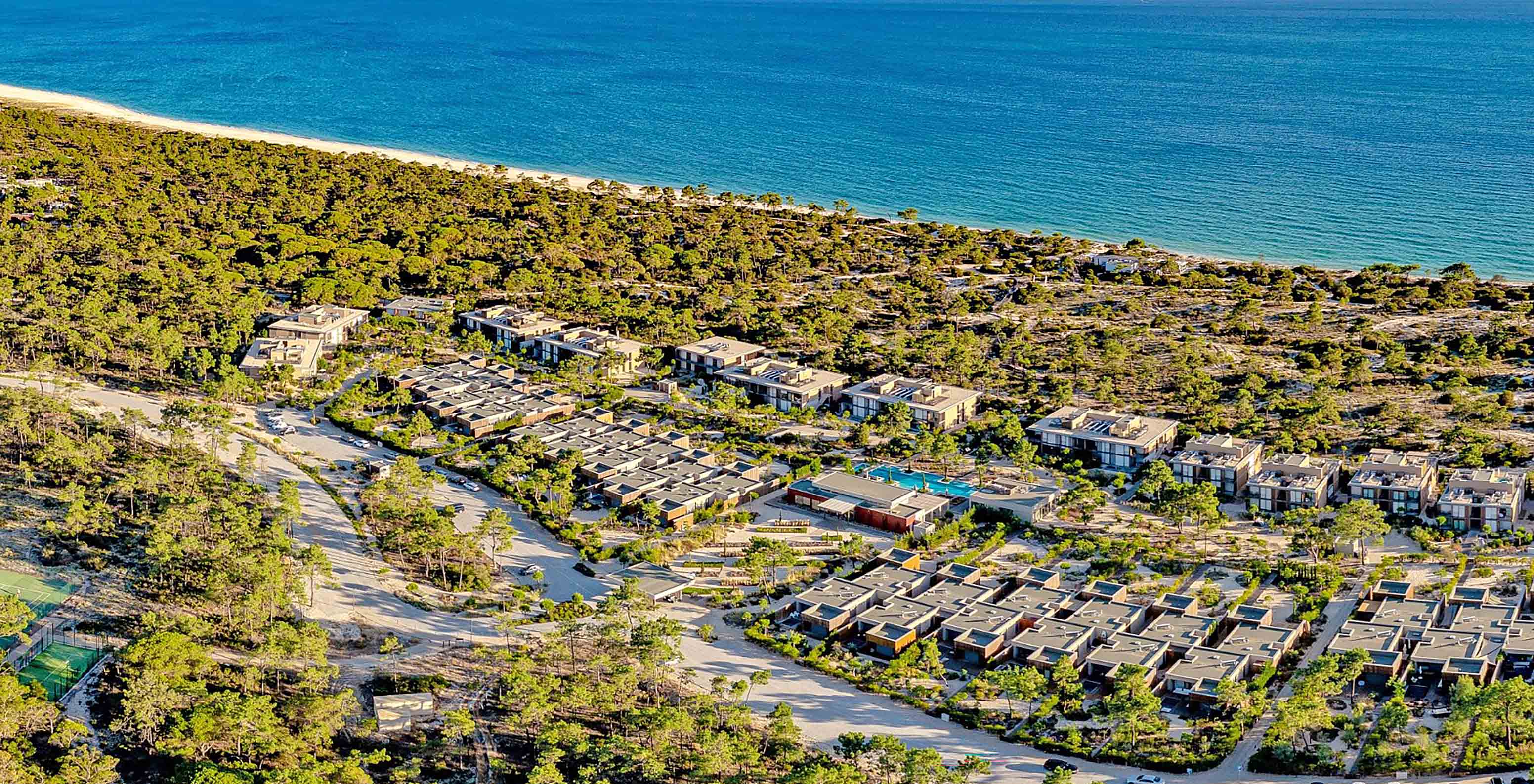 Vista aérea para o Pestana Tróia Eco Resort, perto da praia, com muita vegetação pelo meio e várias casas