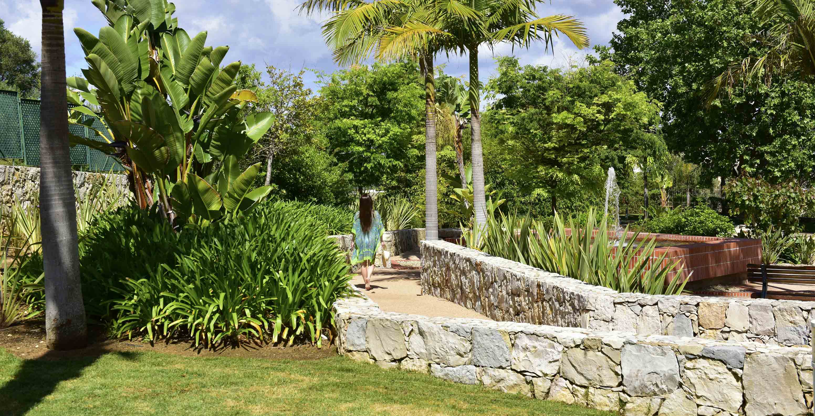 Jardim com árvores e palmeiras, com uma senhora sentada em pedras grandes, com uma fonte rodeada por tijolos