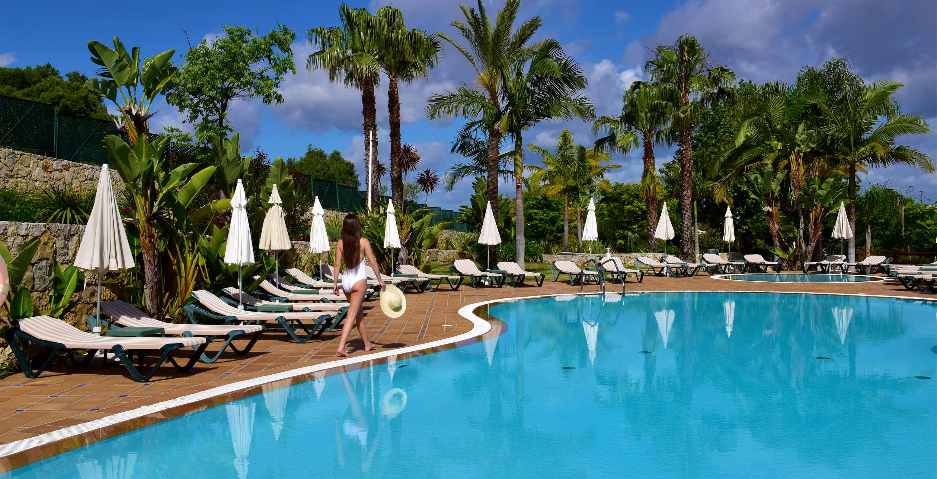 Piscinas exteriores com espreguiçadeiras, chapéus de sol e palmeiras, com uma senhora a andar com fato de banho branco