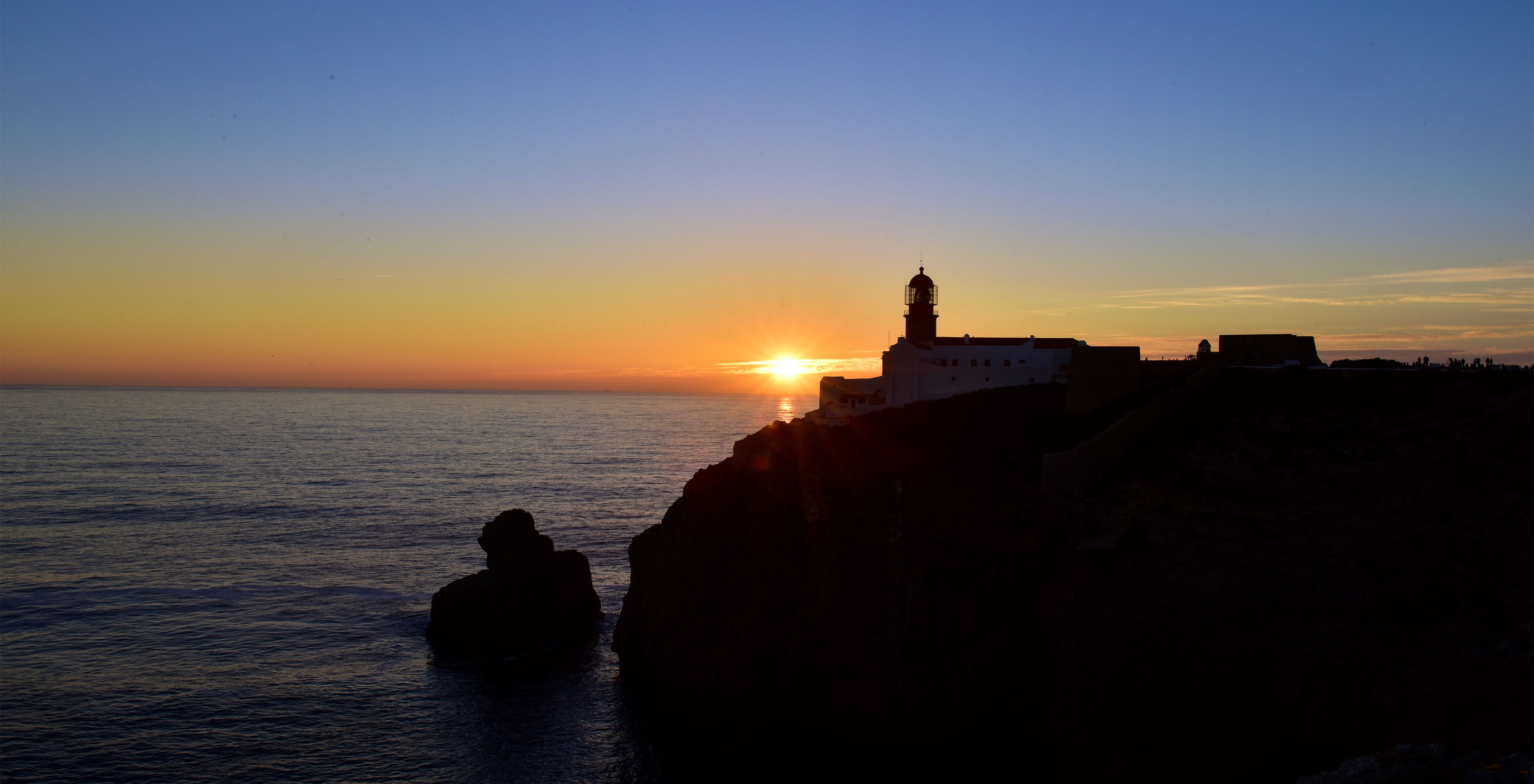 Pôr do Sol numa falésia com o horizonte laranja e um farol no cimo da falésia