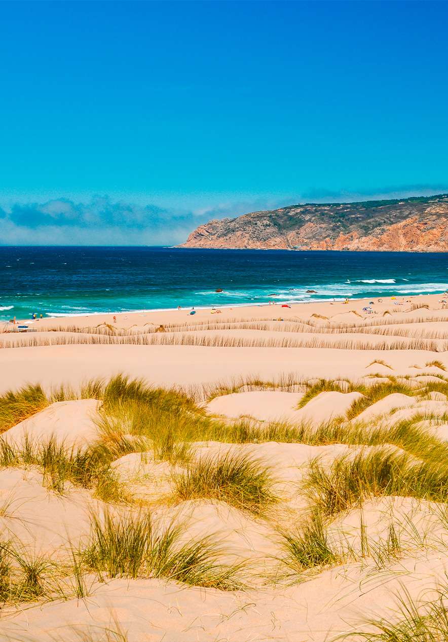 Praia do Guincho em Cascais, onde se vê um dia perfeito de praia, com uma extensão de areia dourada, dunas e mar azul