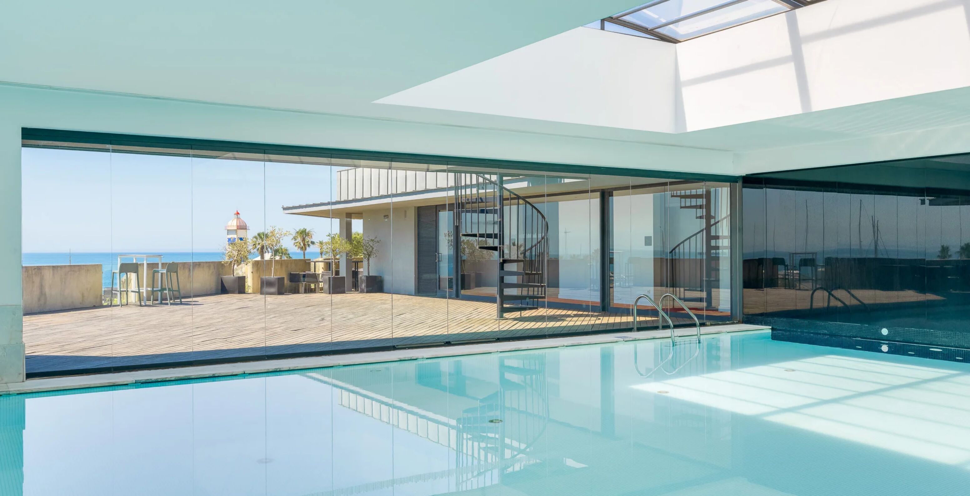Piscina interior com cobertura envidraçada, terraço em madeira e vista marítima junto ao edifício moderno