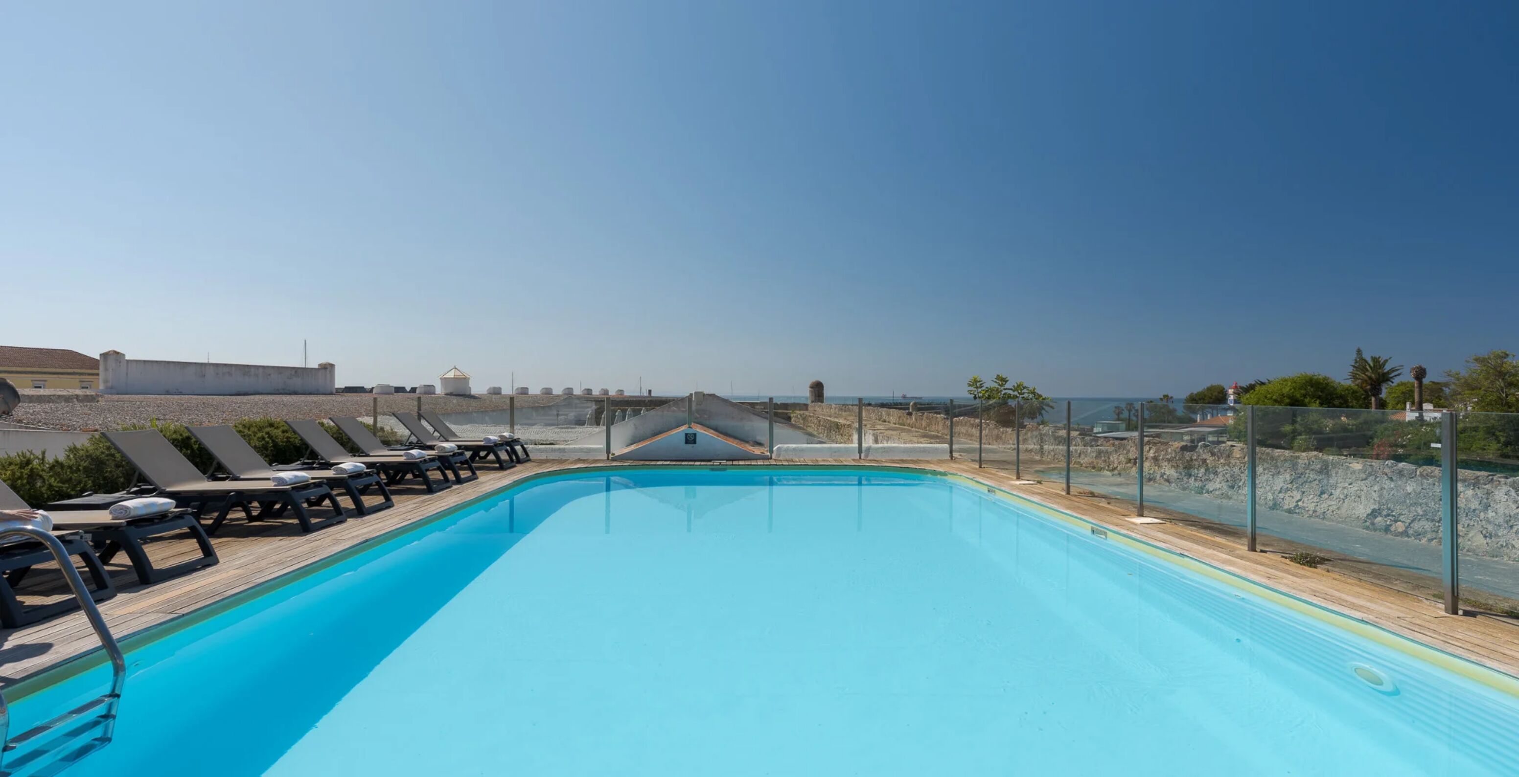 Piscina exterior com espreguiçadeiras junto a um terraço murado e vista ampla para o mar sob céu limpo