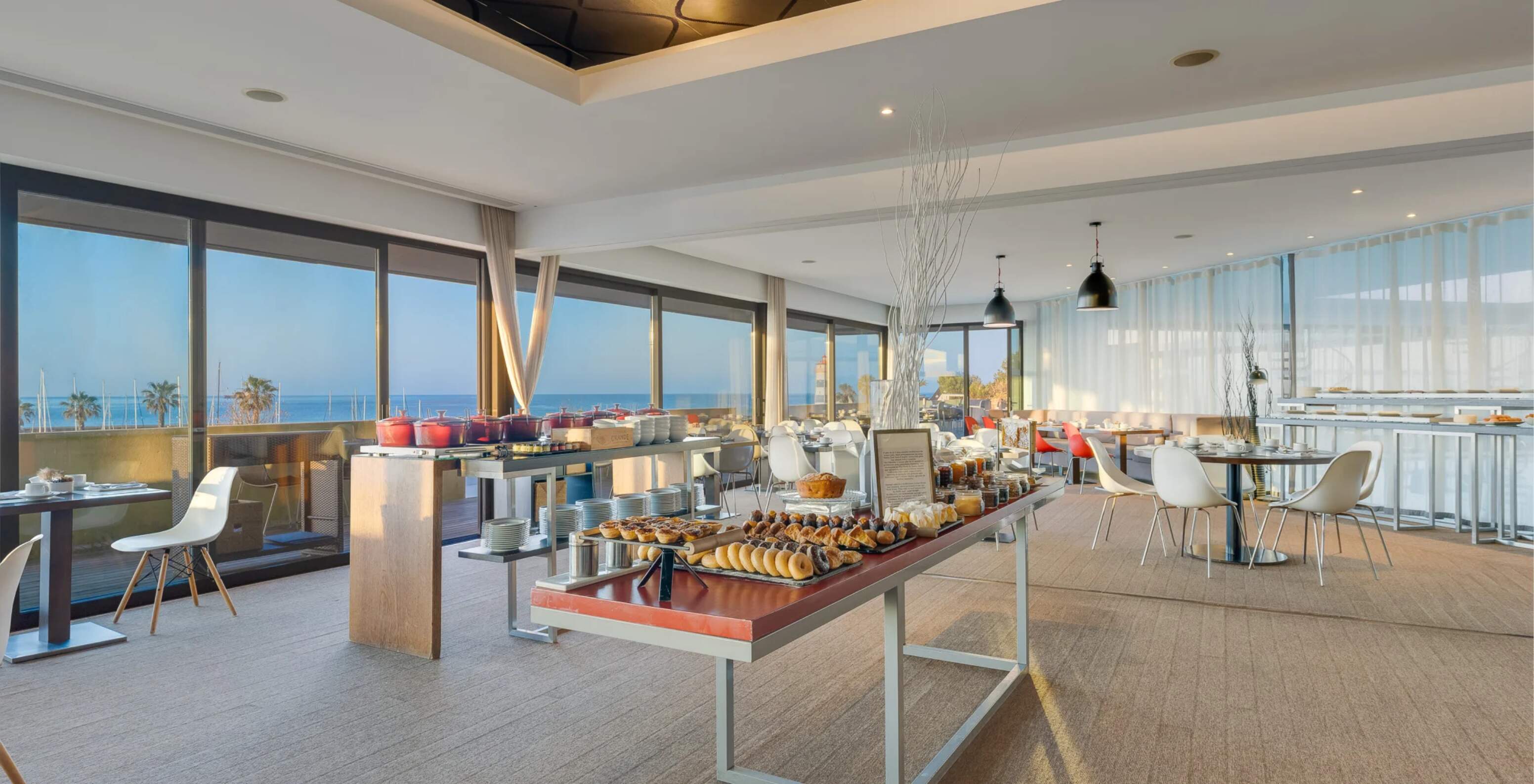 Sala de restaurante luminosa com mesas modernas, buffet de pequeno-almoço e vista mar através das janelas