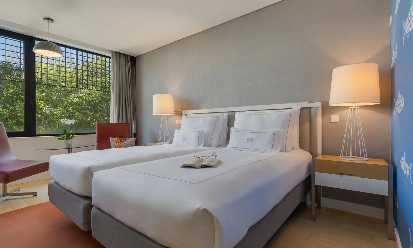 Quarto com duas camas individuais, candeeiros laterais, poltrona vermelha e janela ampla com árvores