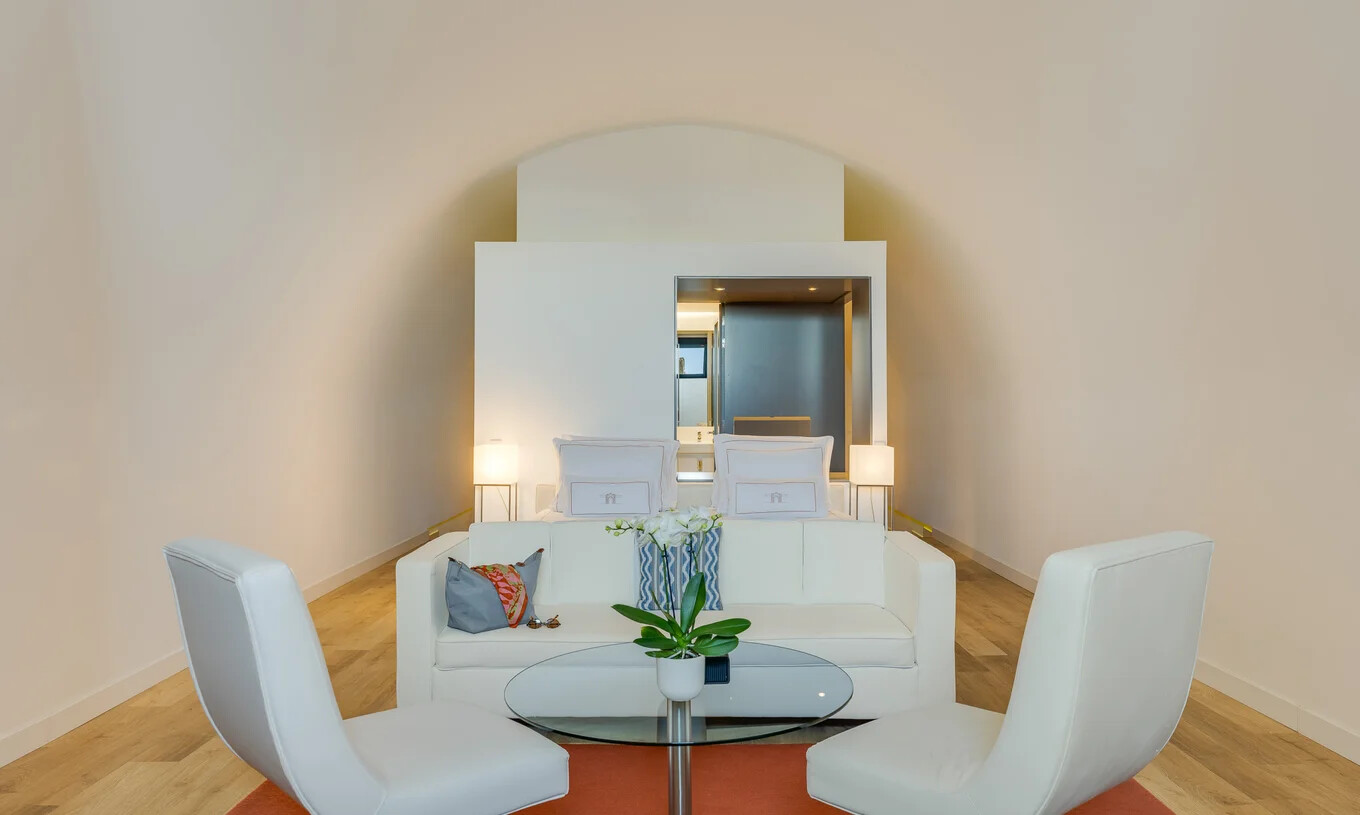 Suite luminosa com sofá branco, duas cadeiras e cama ao fundo junto a mesa redonda com orquídea