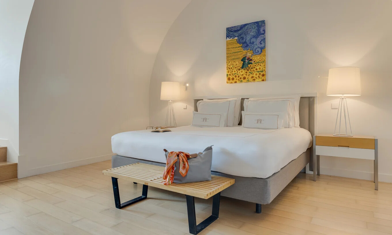 Quarto luminoso com cama de casal, candeeiros laterais, banco em madeira e pintura sobre a cabeceira