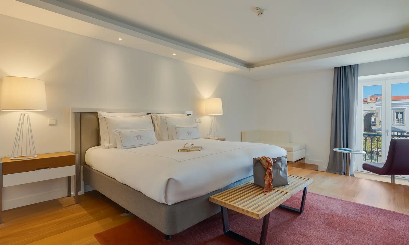 Quarto espaçoso com cama de casal, iluminação suave, tapete vermelho e varanda com vista urbana luminosa