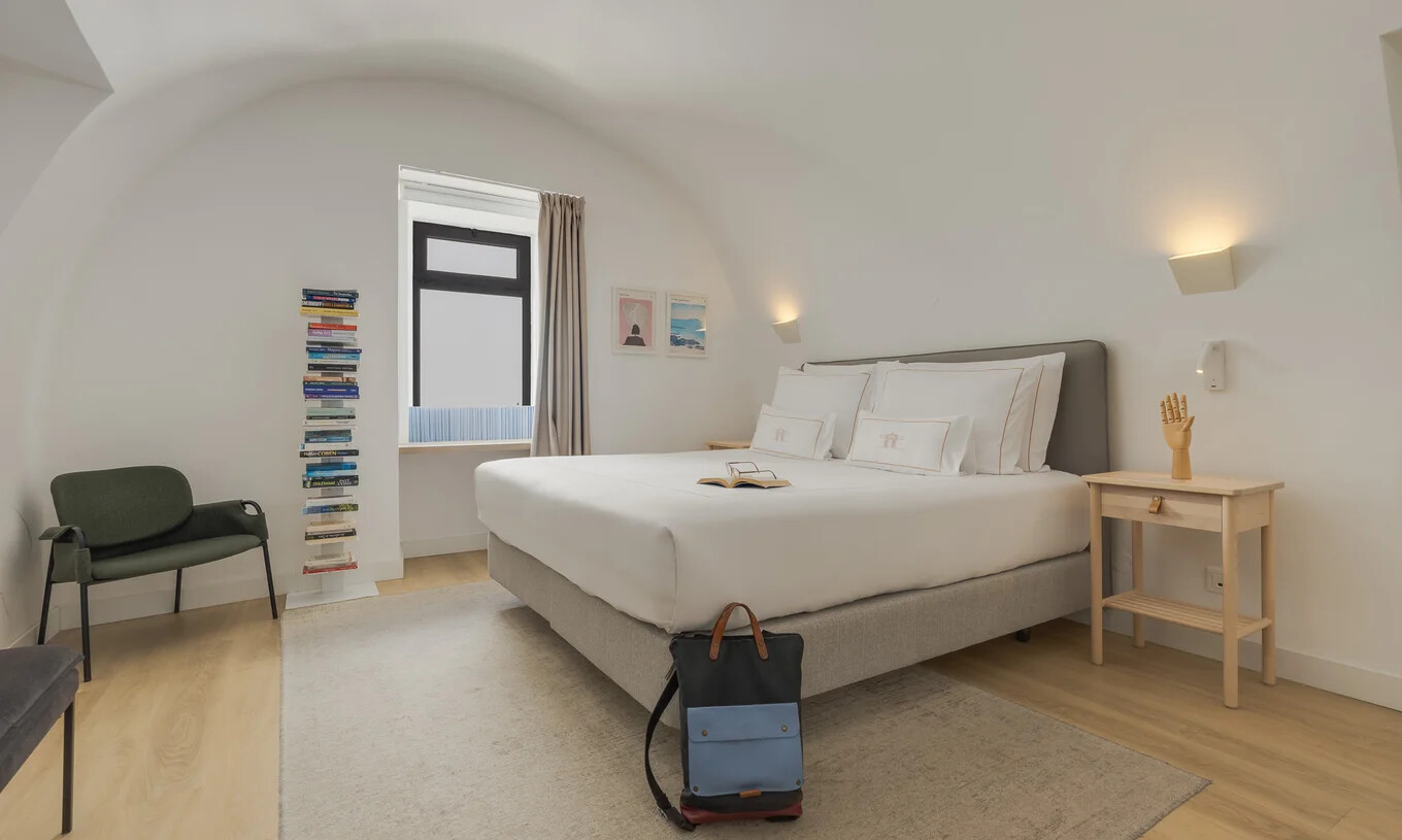 Quarto luminoso com cama de casal, janela com cortinas claras, cadeira preta e mesa de apoio em madeira
